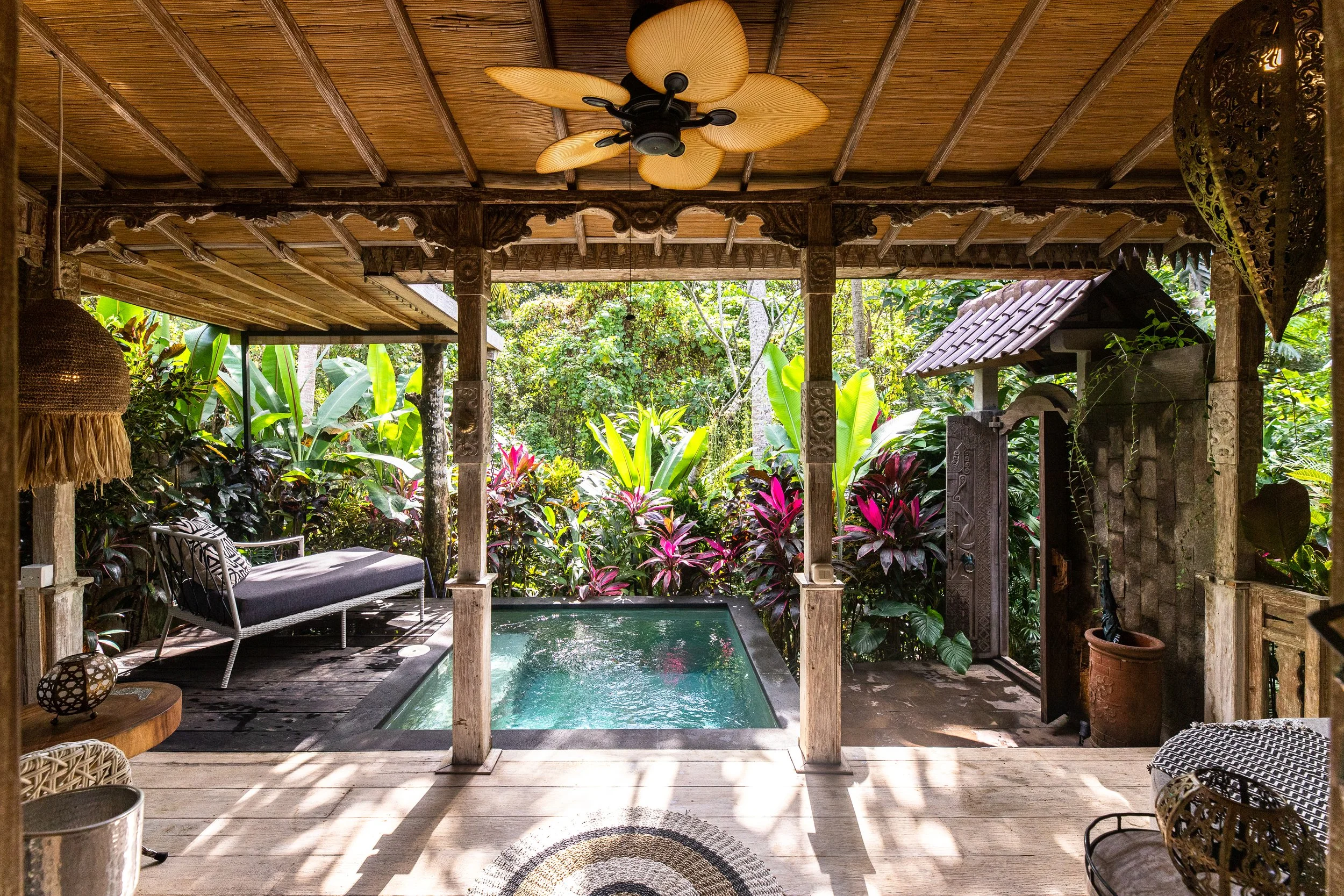 Covered outdoor patio area with a small pool, surrounded by lush tropical plants, and decorated with wicker chairs, cushions, and lanterns, featuring a wooden ceiling fan.