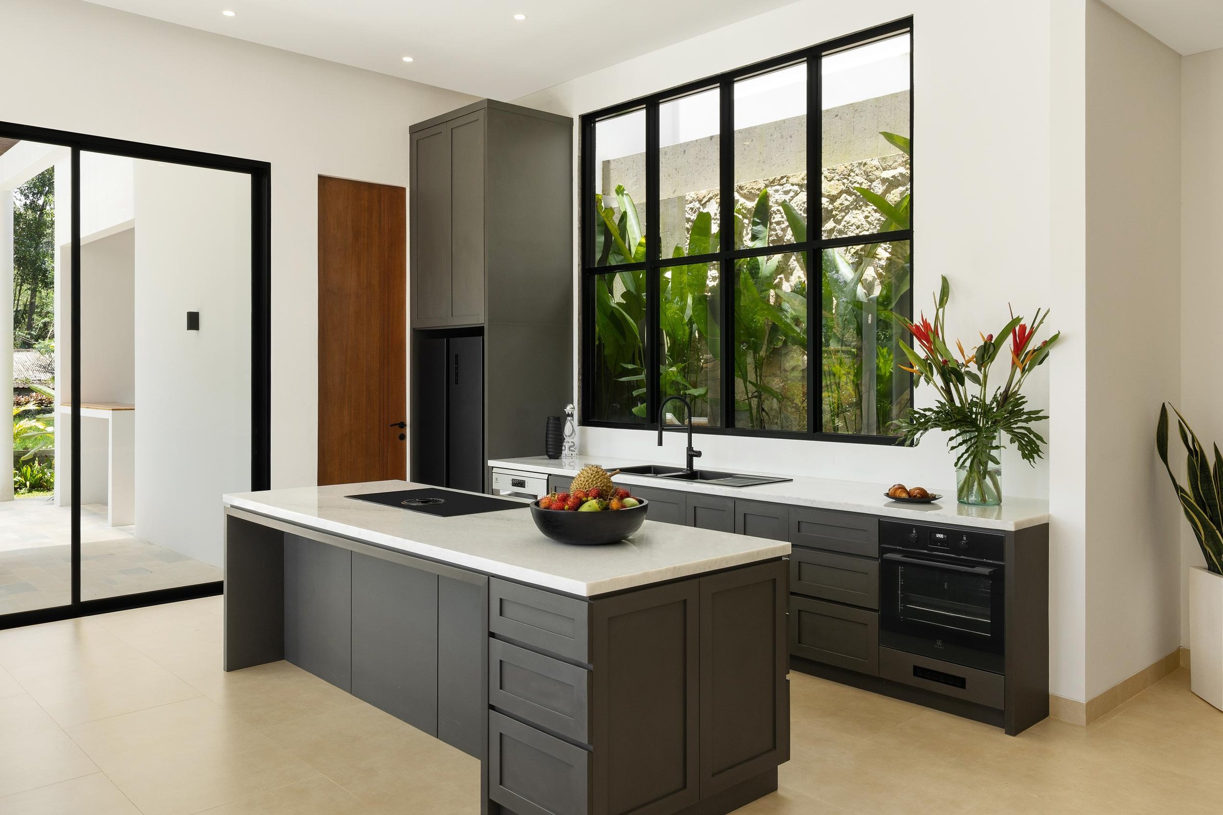 Modern kitchen with large black-framed window, white countertops, and gray cabinets, decorated with a bowl of fruit and plants.