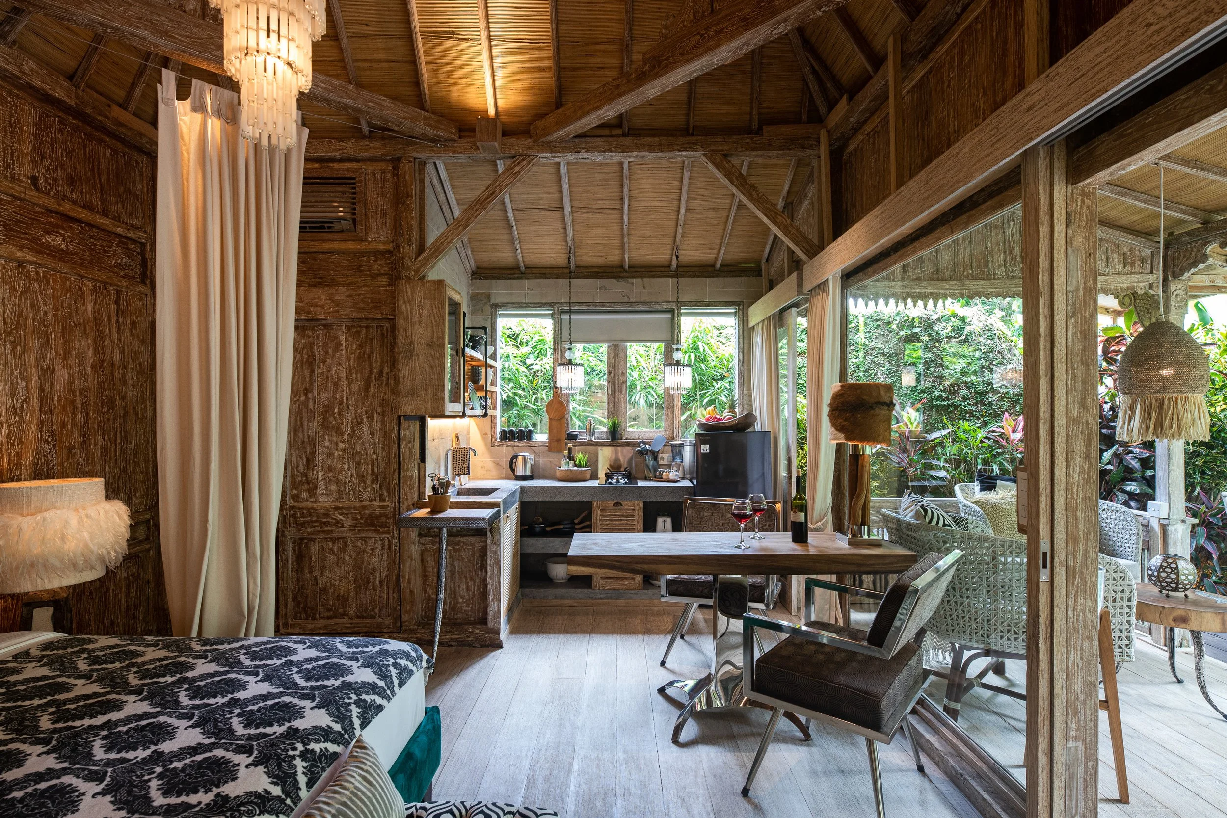 The image shows a cozy, rustic wooden interior of a house or cottage with an open kitchen and dining area, large windows with a view of lush greenery outside, and various chairs and furniture including a table with wine glasses and a bottle on it.