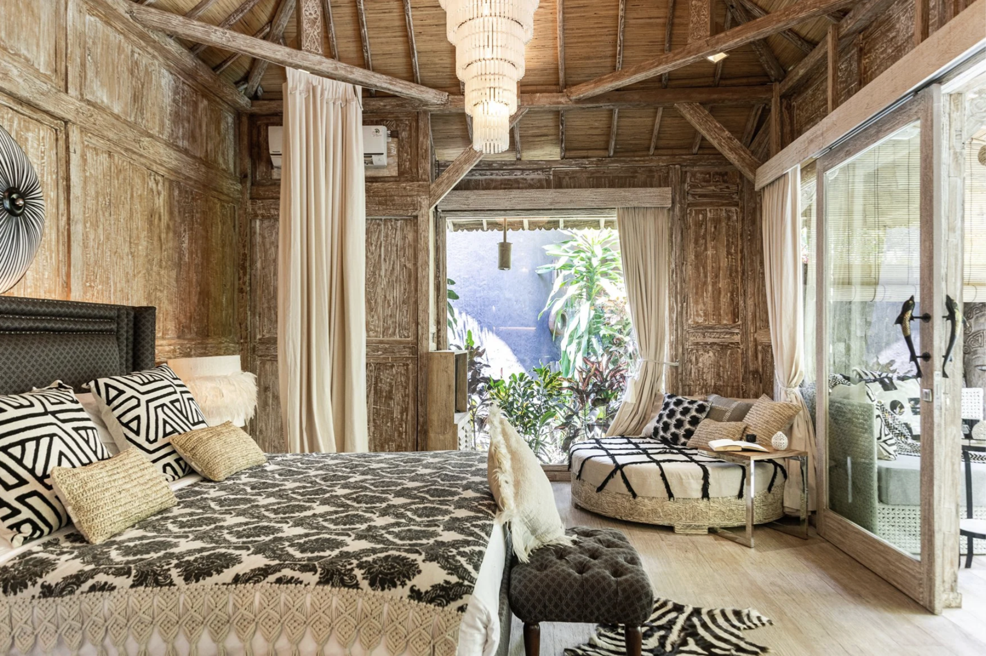 A cozy bedroom with wooden walls and ceiling, a bed with patterned pillows, a round seating area with cushions, and large windows with curtains overlooking a garden with lush green plants.