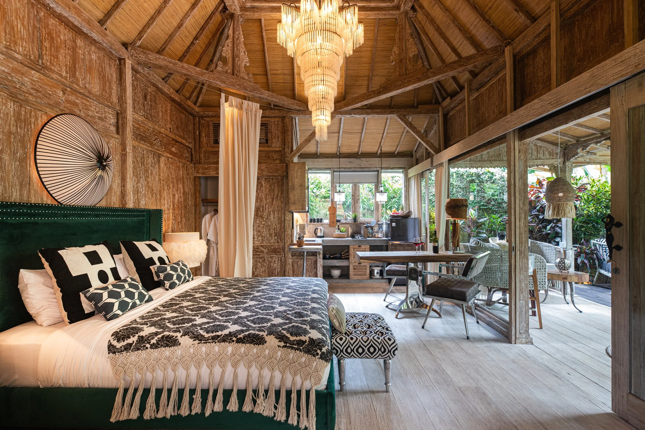 A cozy bedroom with a large bed, black and white patterned pillows, and sheepskin blanket. The room features wooden walls and a high, wooden vaulted ceiling with a large chandelier. There is an open kitchen area and a dining space with outdoor view g
