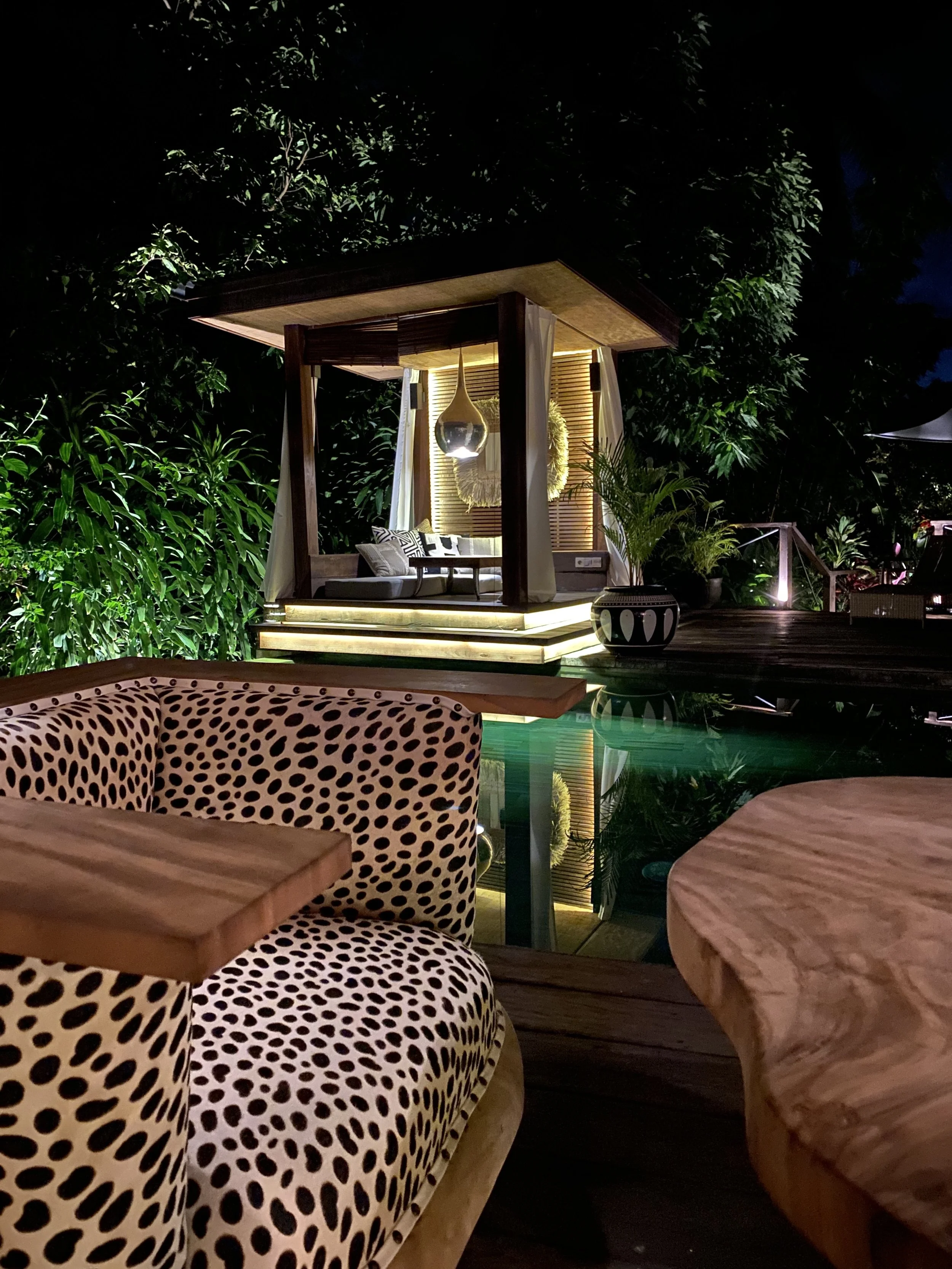 Outdoor seating area with a cozy canopy bed, lighting, and tropical plants at night.