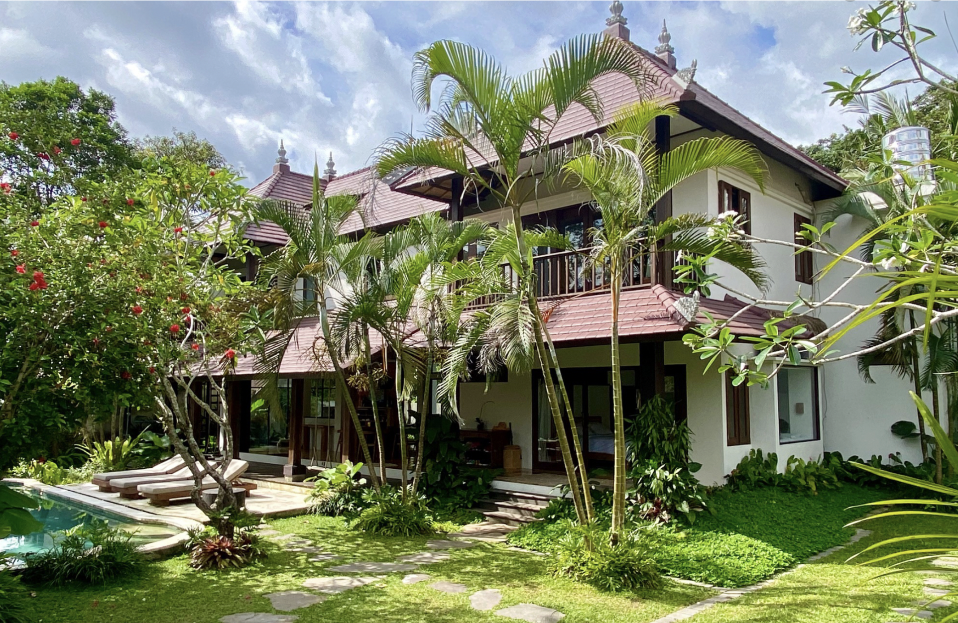 A lush tropical garden with various green plants and trees surrounds a two-story house with a red-tiled roof. The house has large windows and an outdoor balcony. A stone pathway leads through the greenery, and a swimming pool is visible in the foreground.