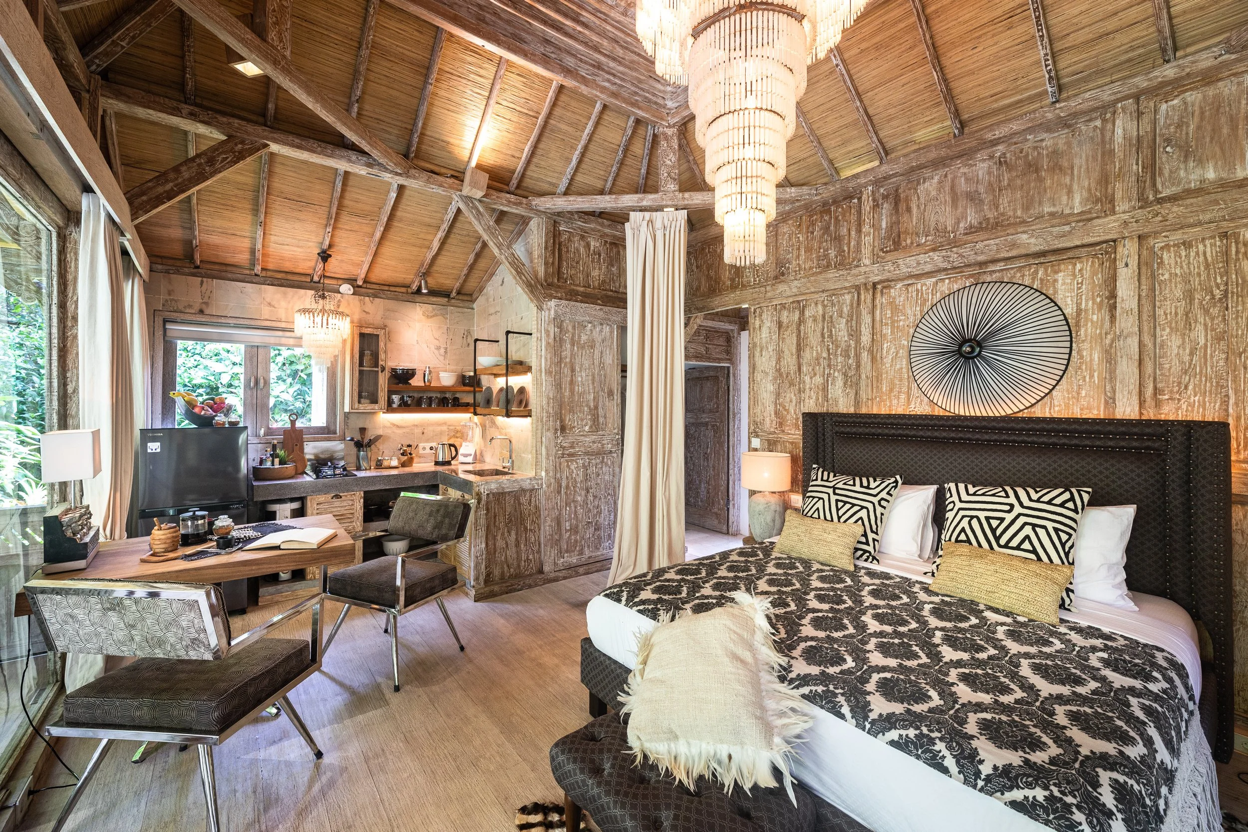 A cozy bedroom with a rustic stone wall, wooden ceiling with exposed beams, and a large chandelier. The bed has black-and-white patterned bedding with pillows, and there is a side table with a lamp. A circular wall art piece decorates the wall behind