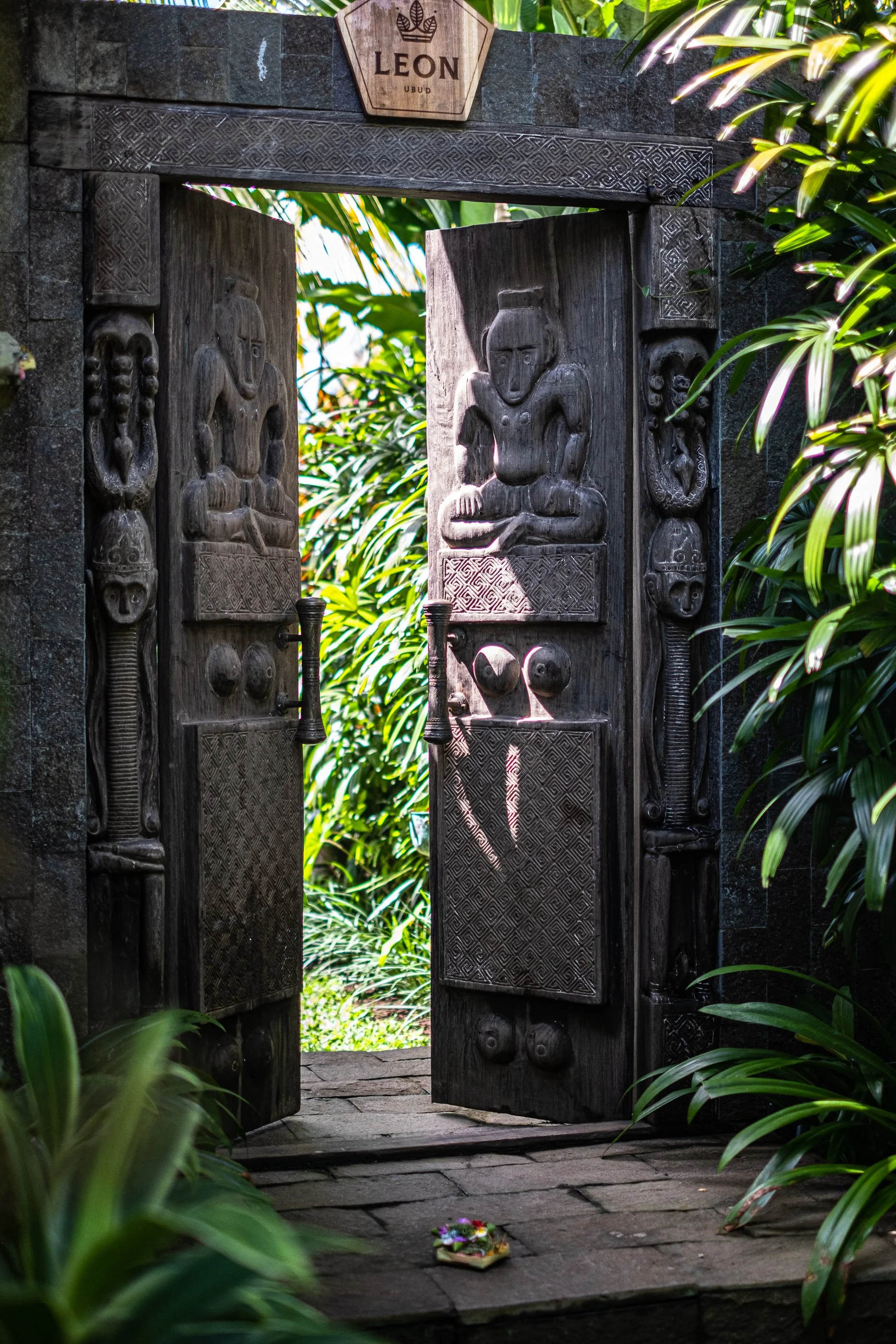 Open wooden door with carvings, leading to a lush green tropical garden. A small floral offering is placed on the stone pathway in front of the door. The sign above the door reads 'LEON UBUD'.