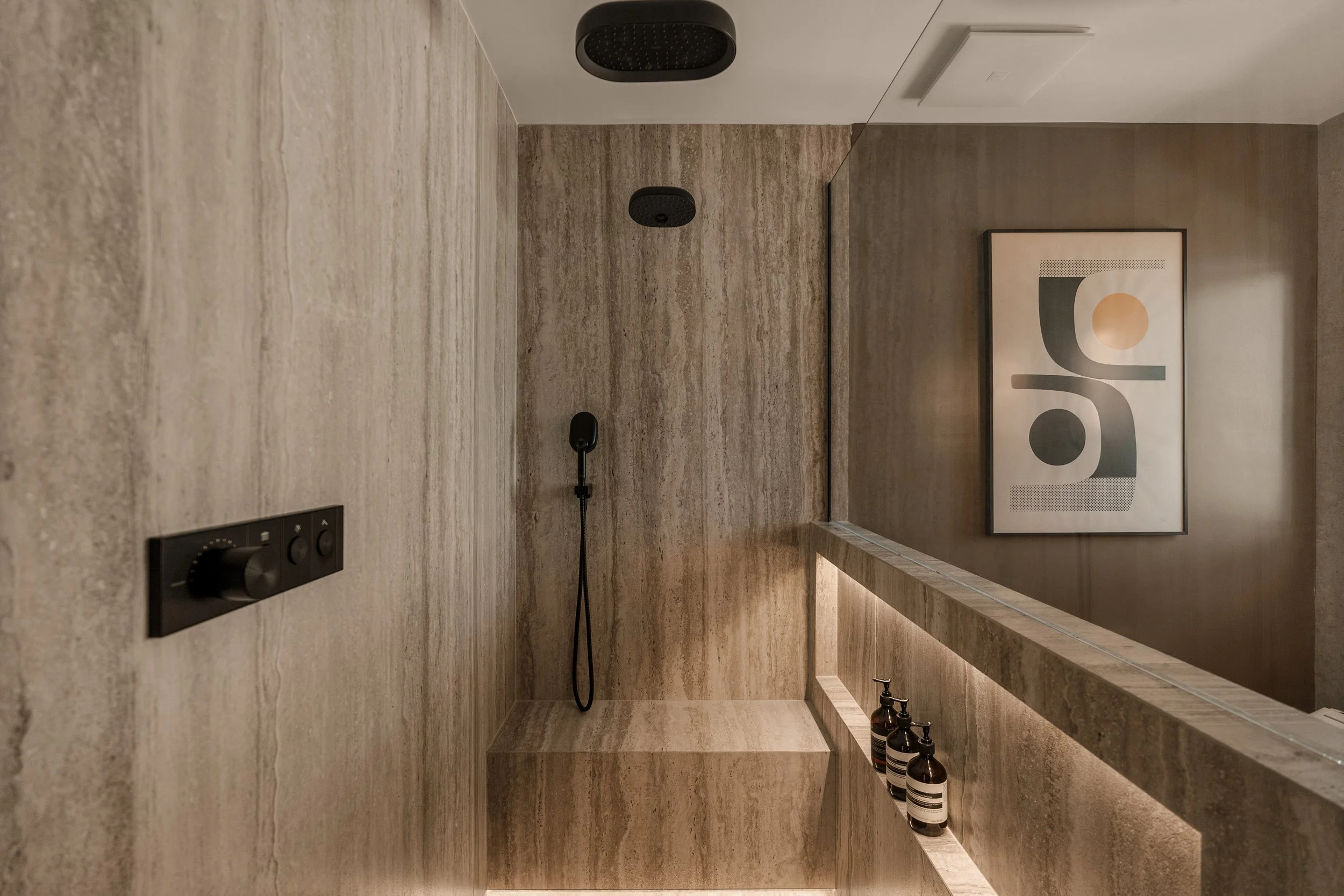 Architectural detail of the spacious walk-in shower with travertine walls, built-in bench seating, and matte black fixtures.