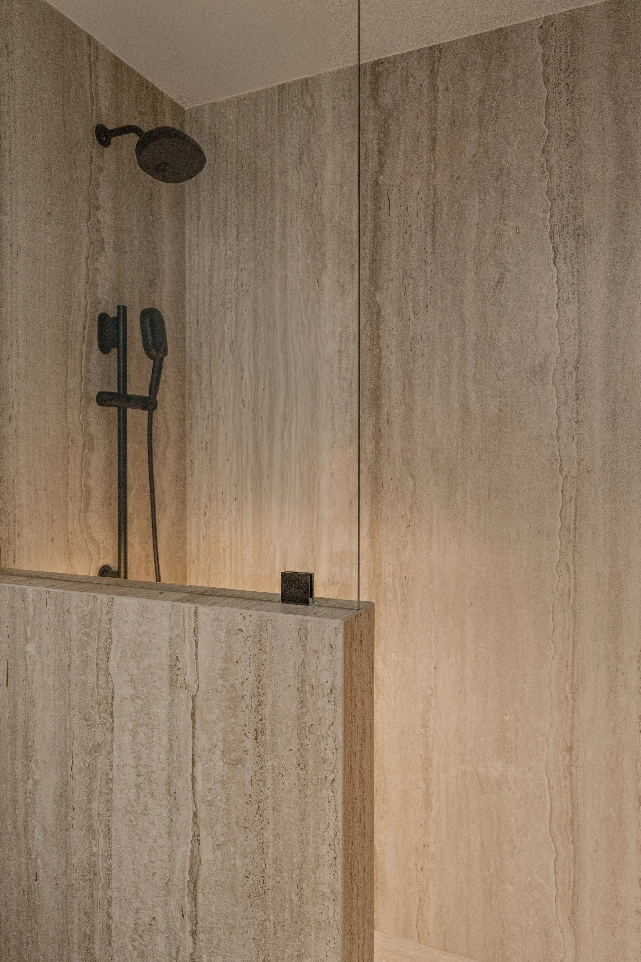 Close-up of the high-end shower system featuring modern matte-black fixtures and travertine wall detail.