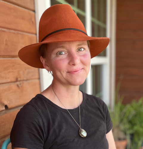 Woman wearing a large brown hat, black shirt, and a pendant necklace, sitting outdoors near a wooden wall and window.