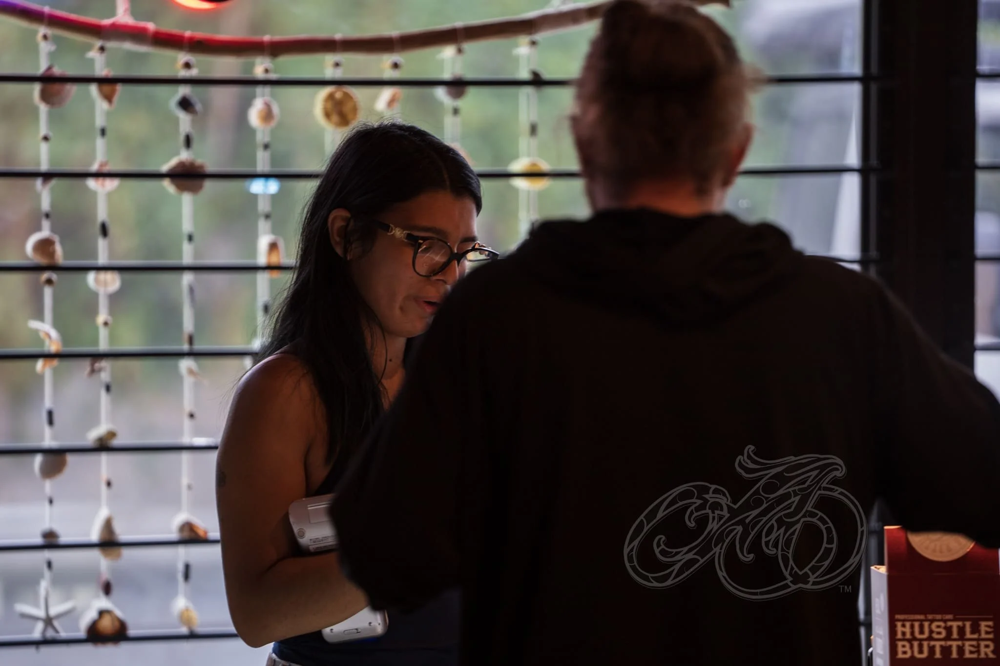 Una mujer de pelo largo y oscuro y gafas sostiene un teléfono móvil y habla con un hombre de pelo corto y sudadera negra con capucha, sentado en un mostrador cerca de una ventana decorada con conchas colgantes.