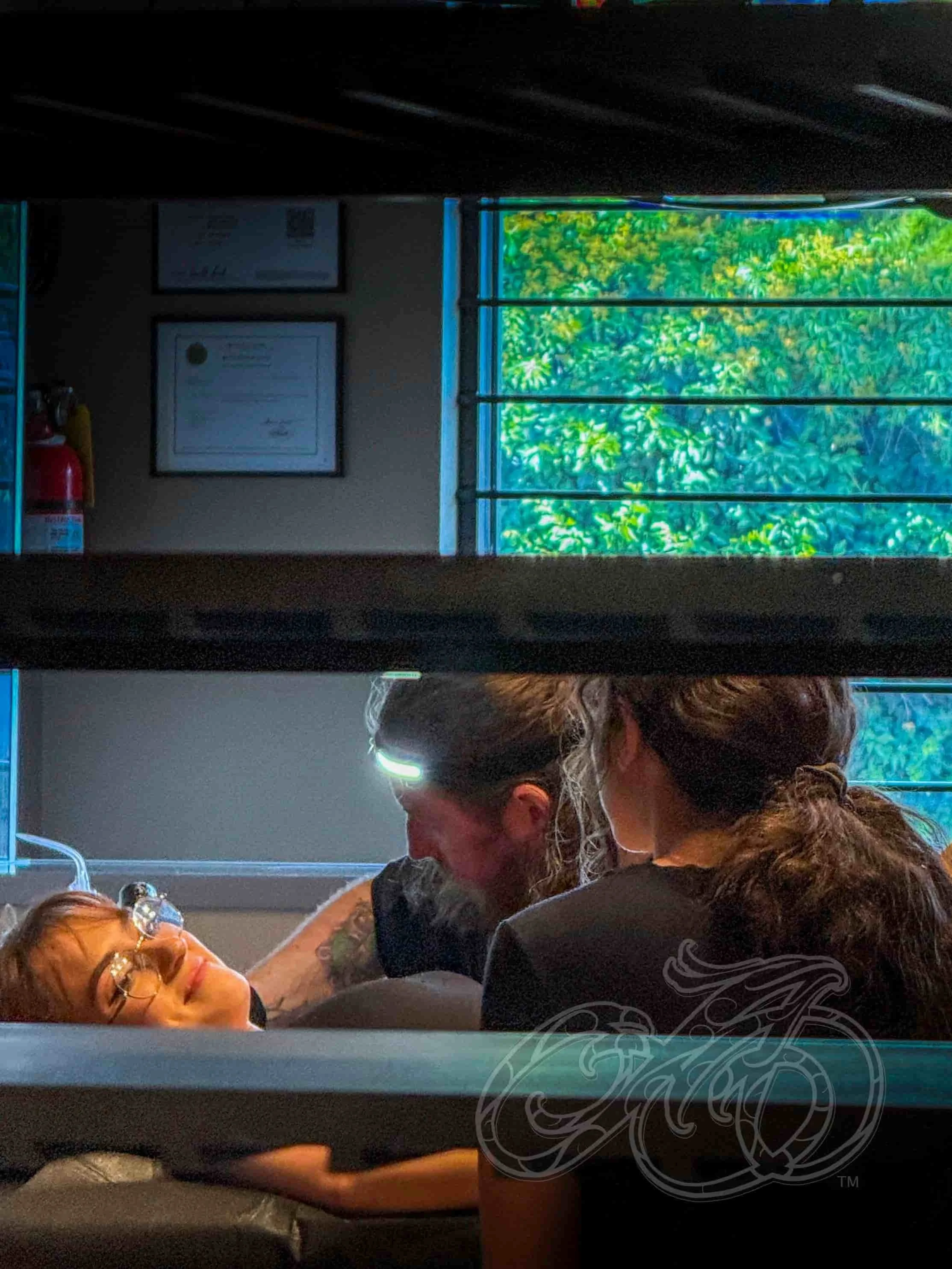Una mujer con gafas tumbada en una mesa, sonríe a un artista del tatuaje con pelo largo y tatuajes, que trabaja en su tatuaje, en una habitación con vistas a una ventana con follaje verde.