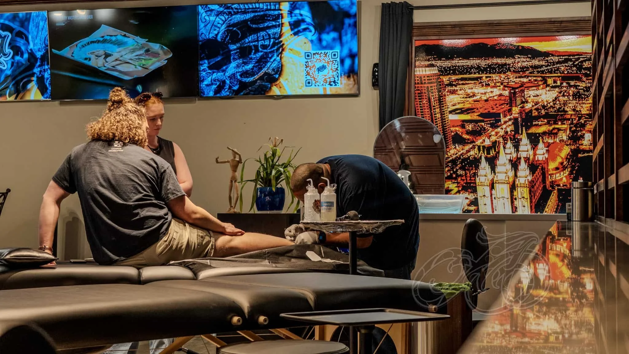 Una persona se hace un tatuaje en la pierna por un tatuador en un estudio moderno, con dos mujeres de pie cerca y un gran mural de un paisaje urbano en la pared.