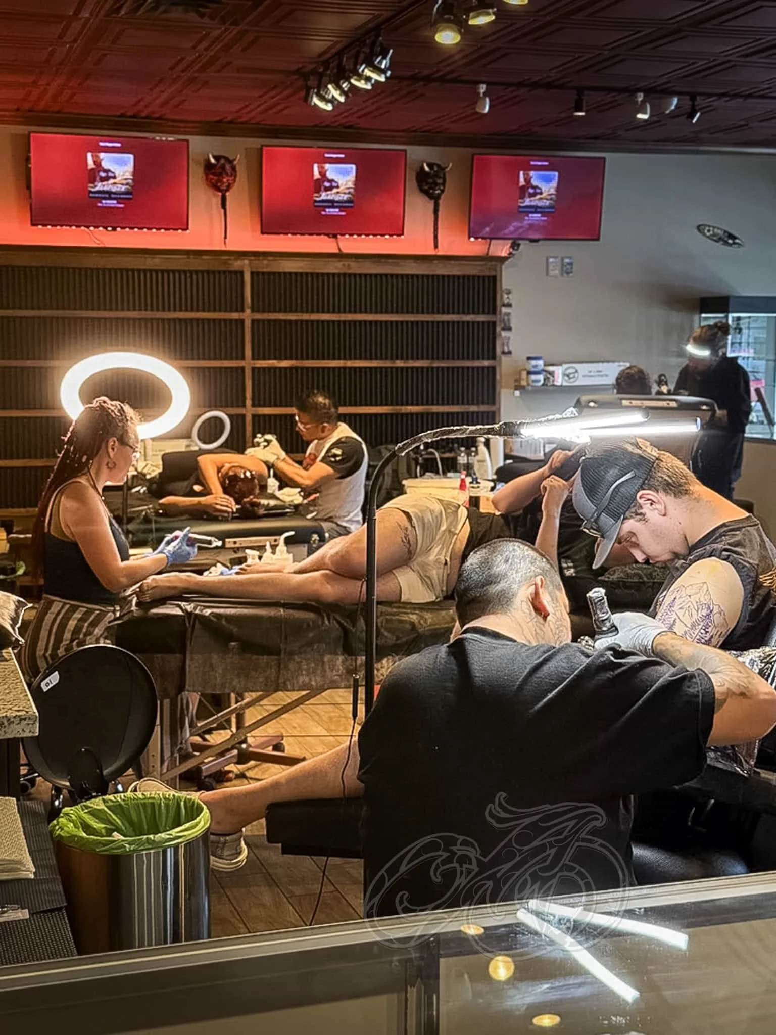 Artistas tatuadores trabajando con clientes en un estudio de tatuajes, con los clientes tumbados en las mesas y los artistas tatuadores concentrados en su trabajo, iluminados por luces cenitales.
