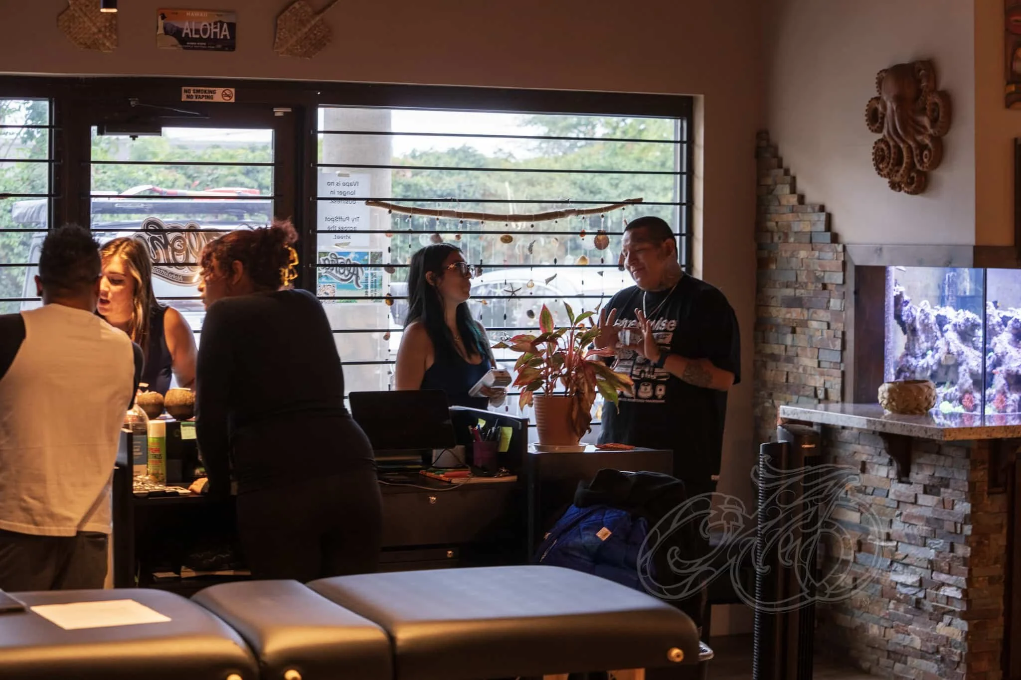 Un grupo de personas conversa en el interior de un restaurante, con una ventana detrás que muestra los árboles del exterior. Hay una maceta sobre el mostrador y una pecera empotrada en una pared de ladrillo. El ambiente parece informal y social.