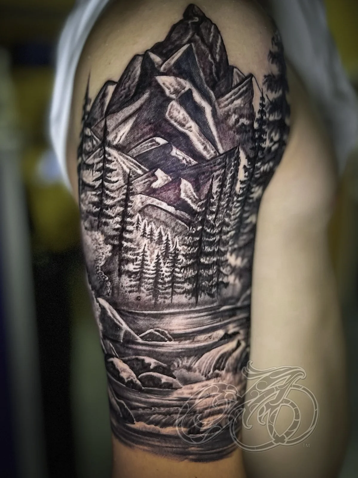 Black and gray realistic tattoo of a mountain landscape with trees, a river, and mountains in the background on a person's upper arm.