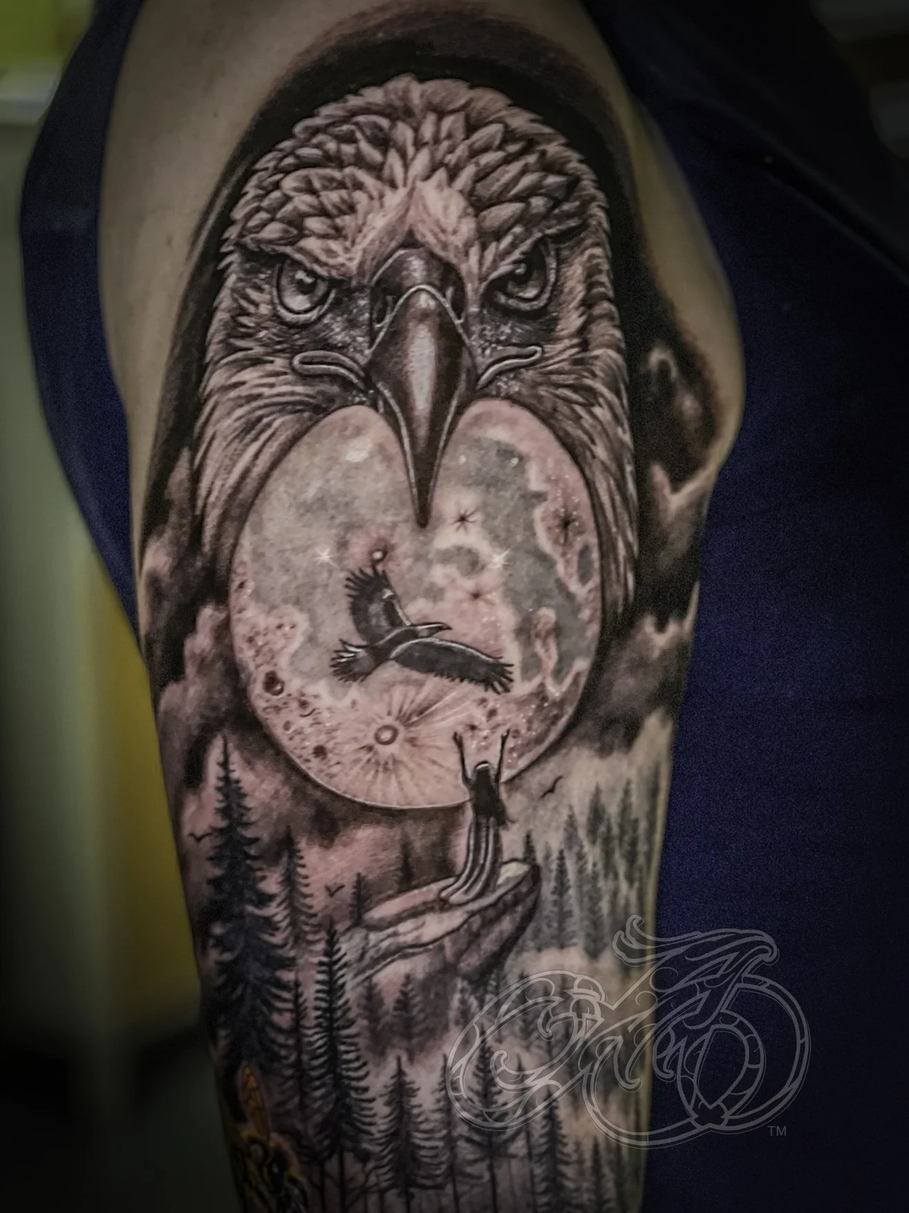 Black and grey tattoo of an eagle's head with an moon in space scene with a person at the edge of a cliff, back turned, looking at the eagle, with pine trees and a starry sky.