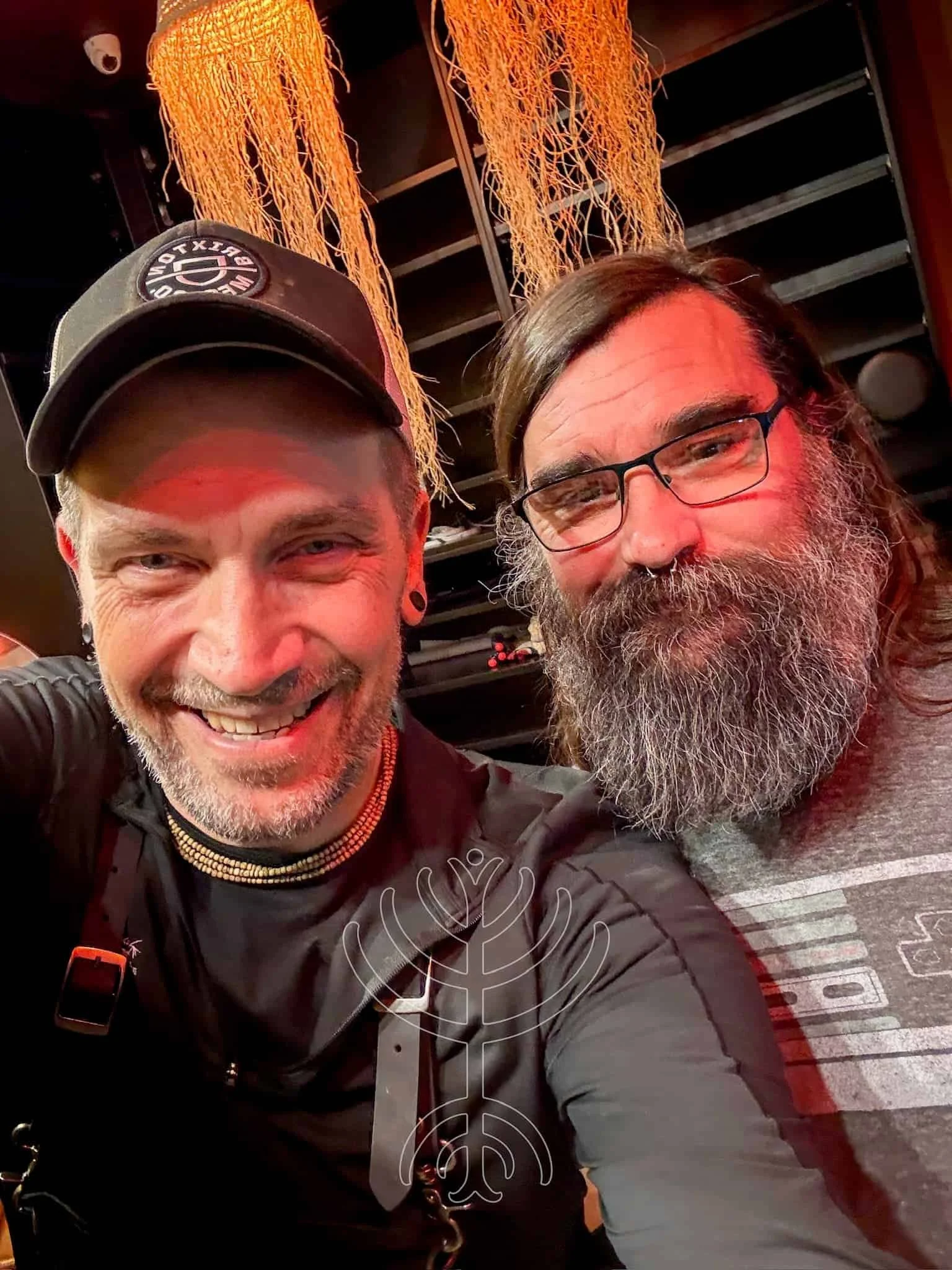 Dos hombres sonrientes se toman un selfie juntos. Uno lleva barba, gafas y camisa gris. El otro lleva perilla, pelo corto, gorra y pendientes. En el fondo cuelgan hilos decorativos de color naranja.
