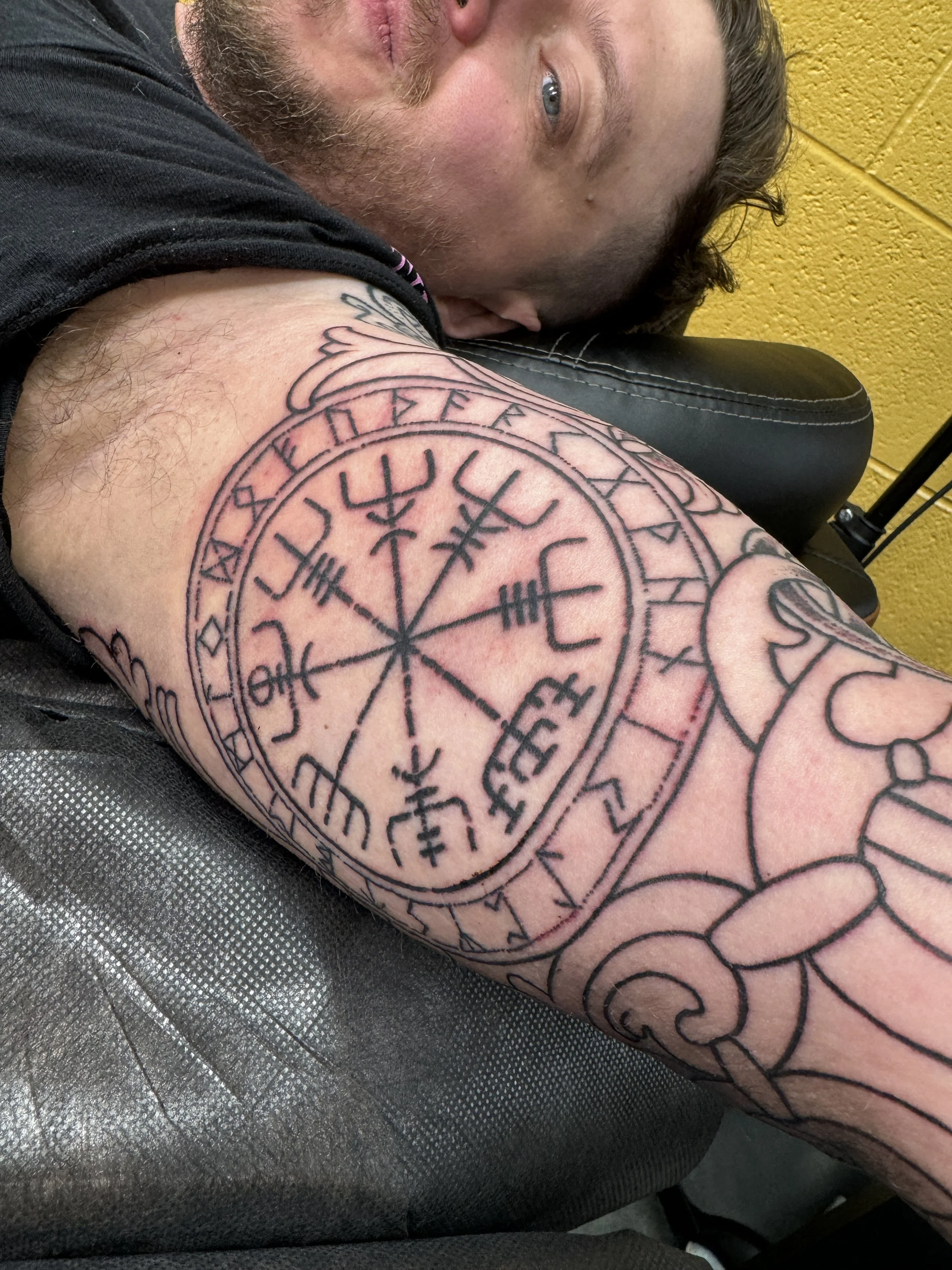 A man with a tattoo of a Norse Vegvisir symbol on his forearm, lying on a chair with a yellow wall in the background.