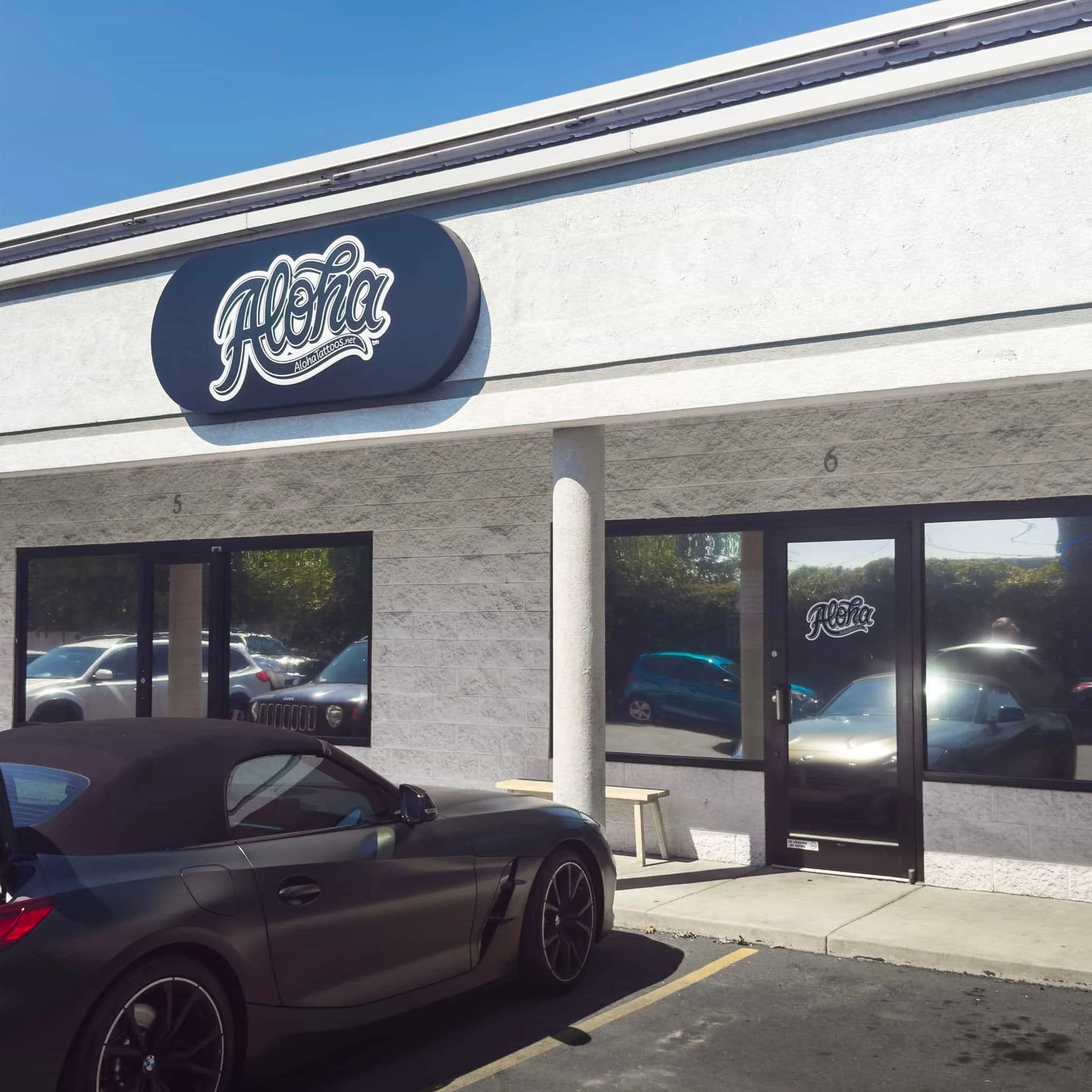 Exterior de una tienda con un letrero que dice "Aloha" y un logotipo. Hay un coche descapotable negro aparcado delante, y varios otros coches aparcados reflejados en las ventanas.