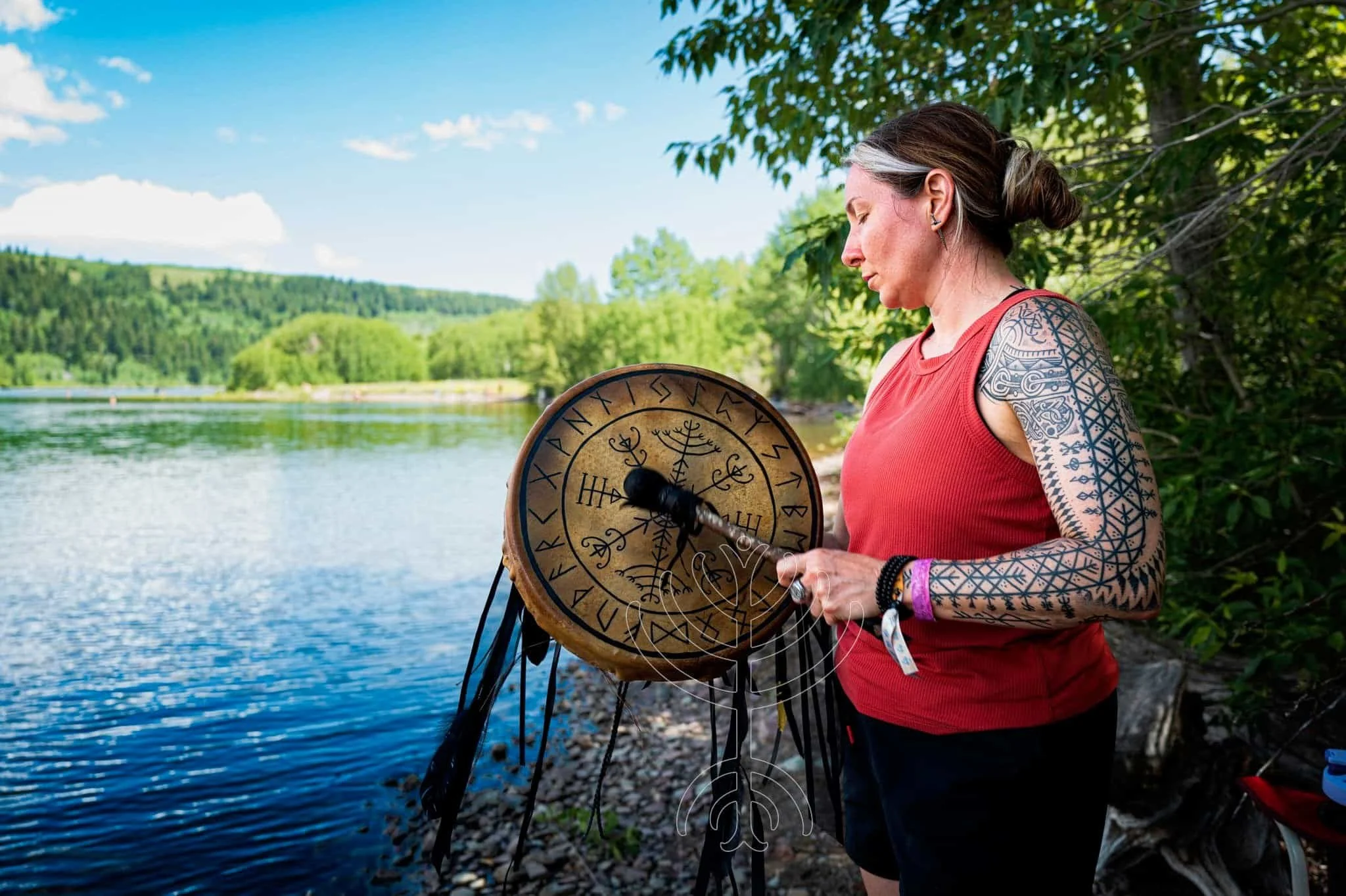 Una mujer con tatuajes en los brazos está junto a un río, tocando un gran tambor con símbolos rúnicos, al aire libre en un día soleado.