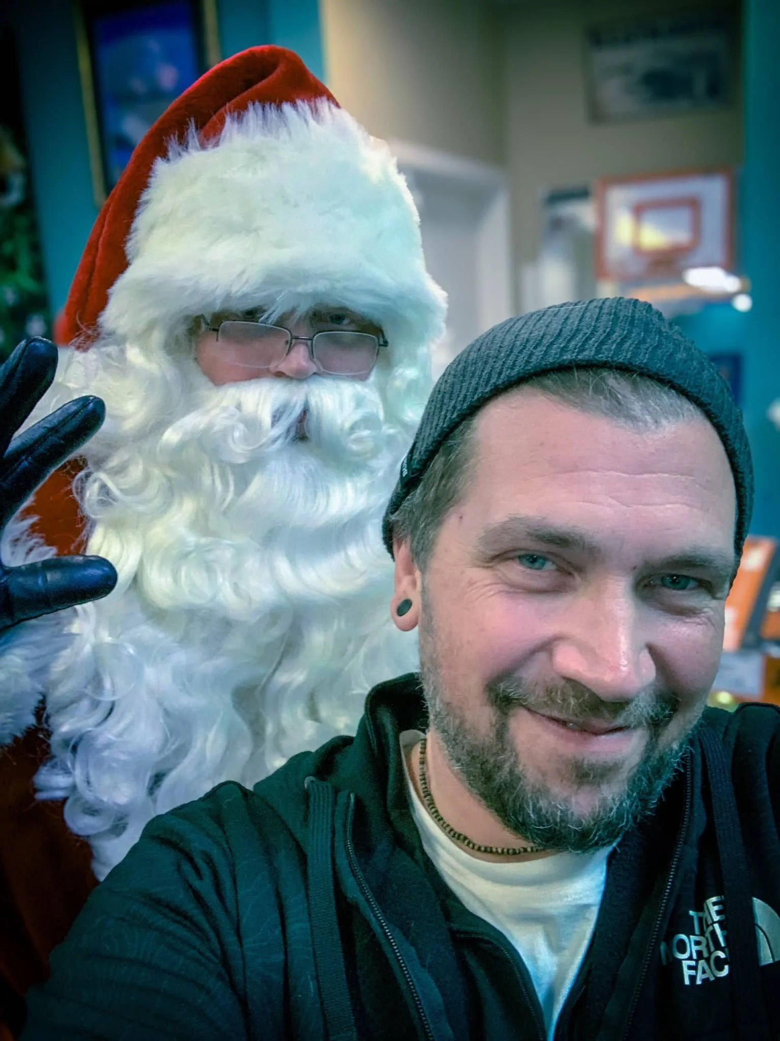 Un hombre se hace un selfie con una persona vestida de Papá Noel. El hombre tiene barba, lleva un gorro y una chaqueta negra y sonríe. Papá Noel lleva gafas, gorro rojo con ribete blanco, barba blanca rizada y hace un gesto con la mano con un guante.