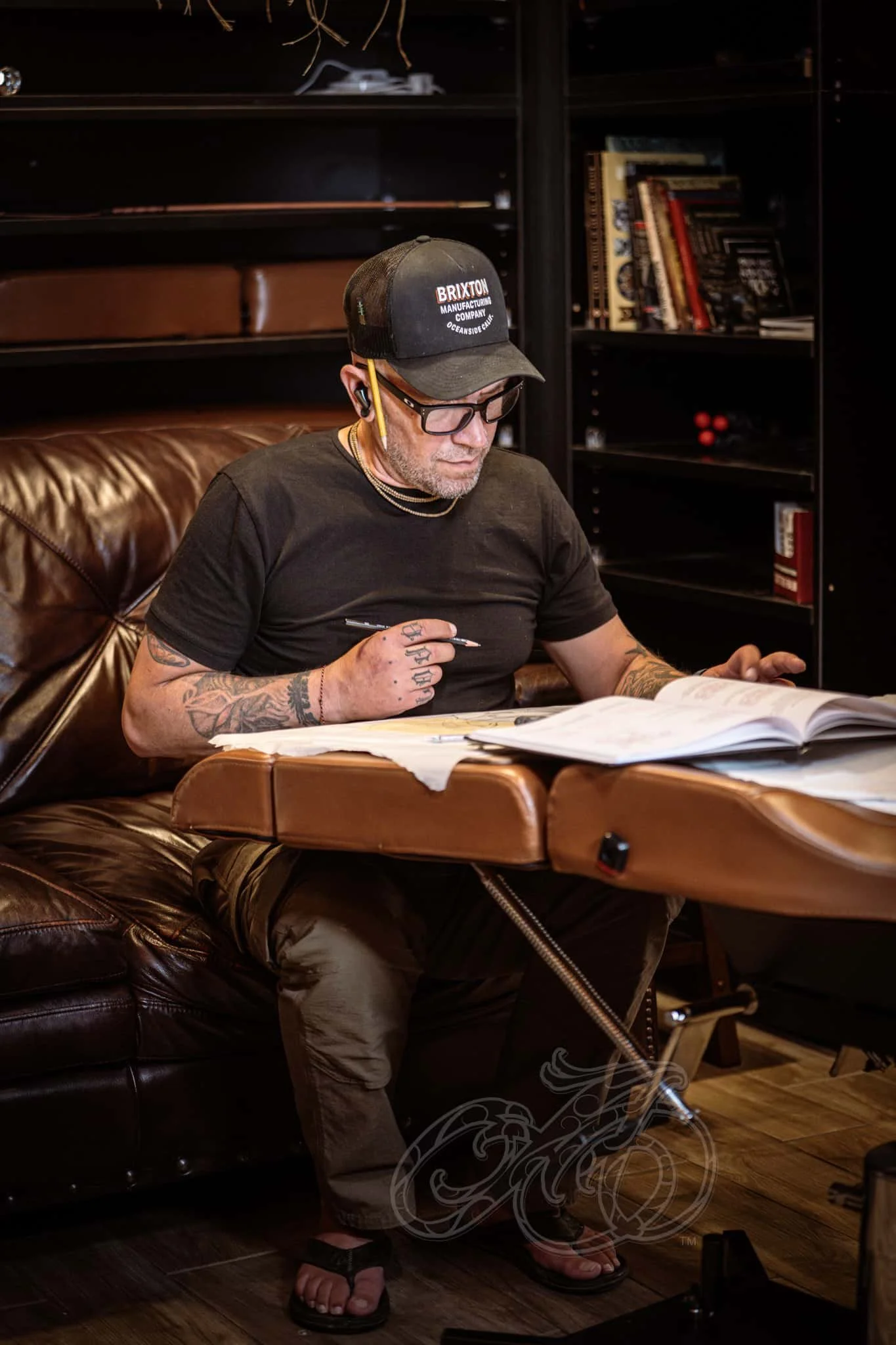 Un hombre con tatuajes, gafas y gorra estudia unos documentos ante una mesa de madera en una acogedora habitación con estanterías oscuras.