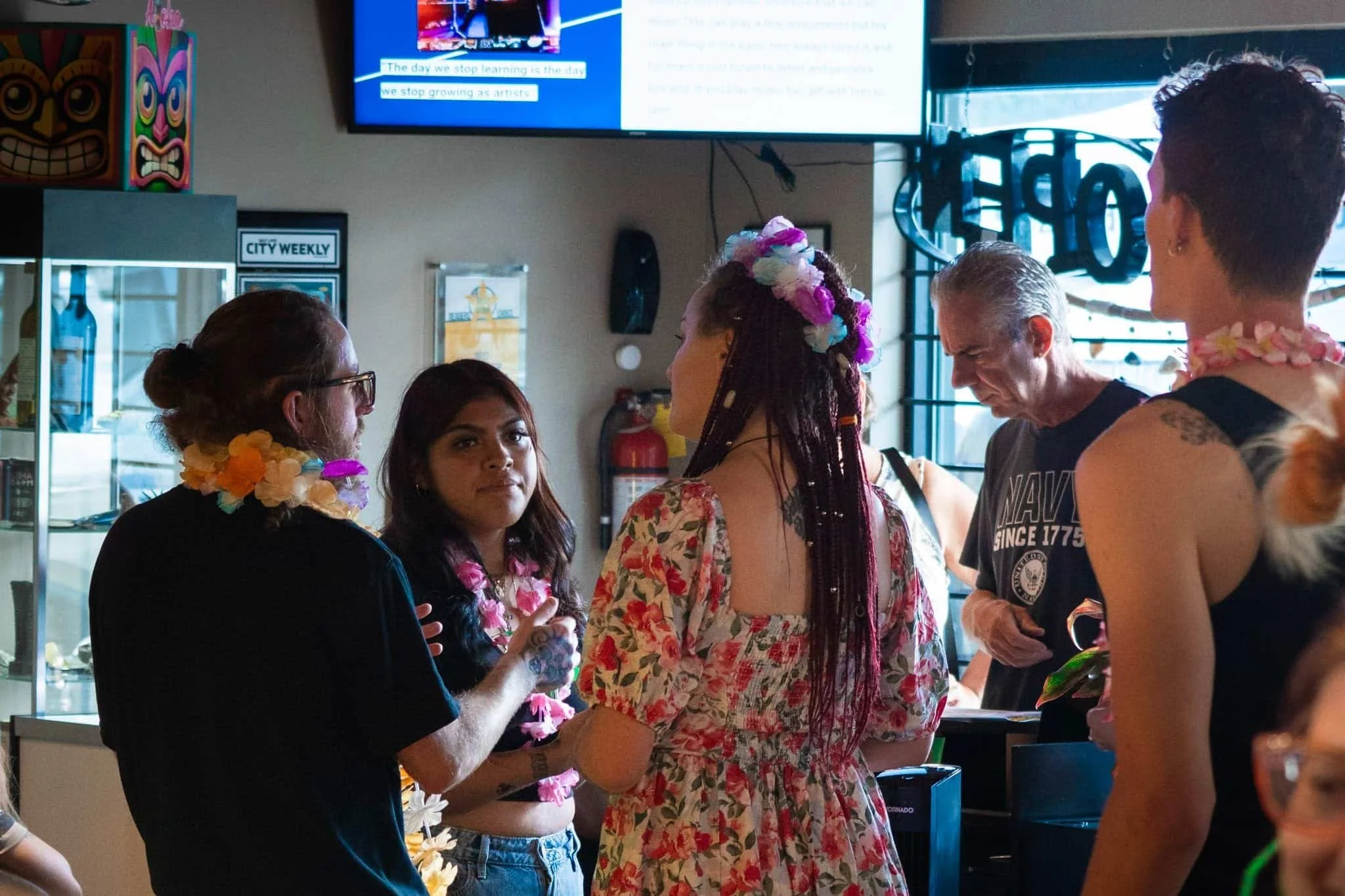 Un grupo de cinco personas reunidas en el interior, algunas con leis y adornos florales, conversan. El ambiente parece informal, con una pantalla de televisión y un letrero de neón al fondo, y una vitrina de cristal a la izquierda.