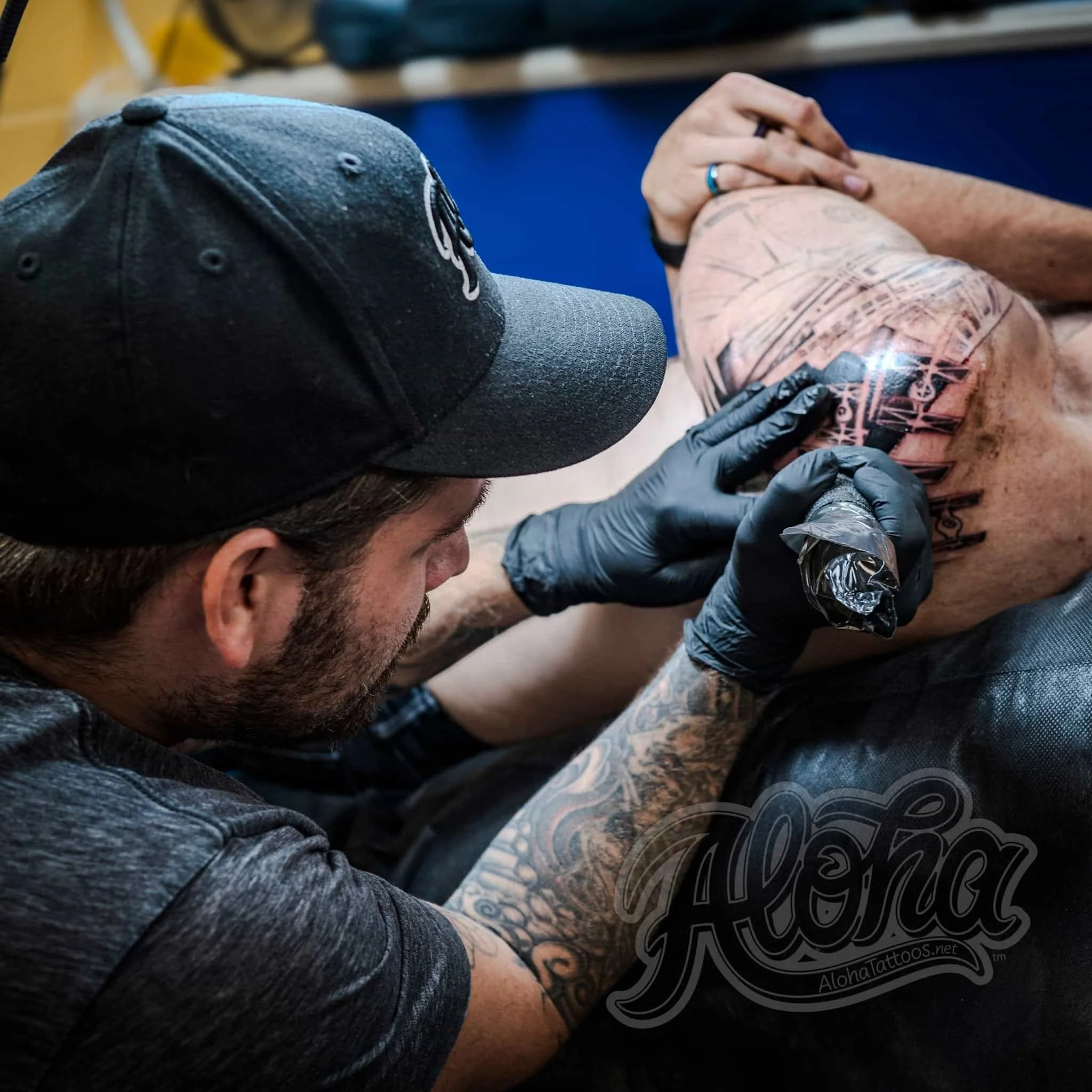 Un tatuador con gorro negro tatúa un gran dibujo en la parte superior de la pierna de un cliente, con tinta negra y guantes negros.