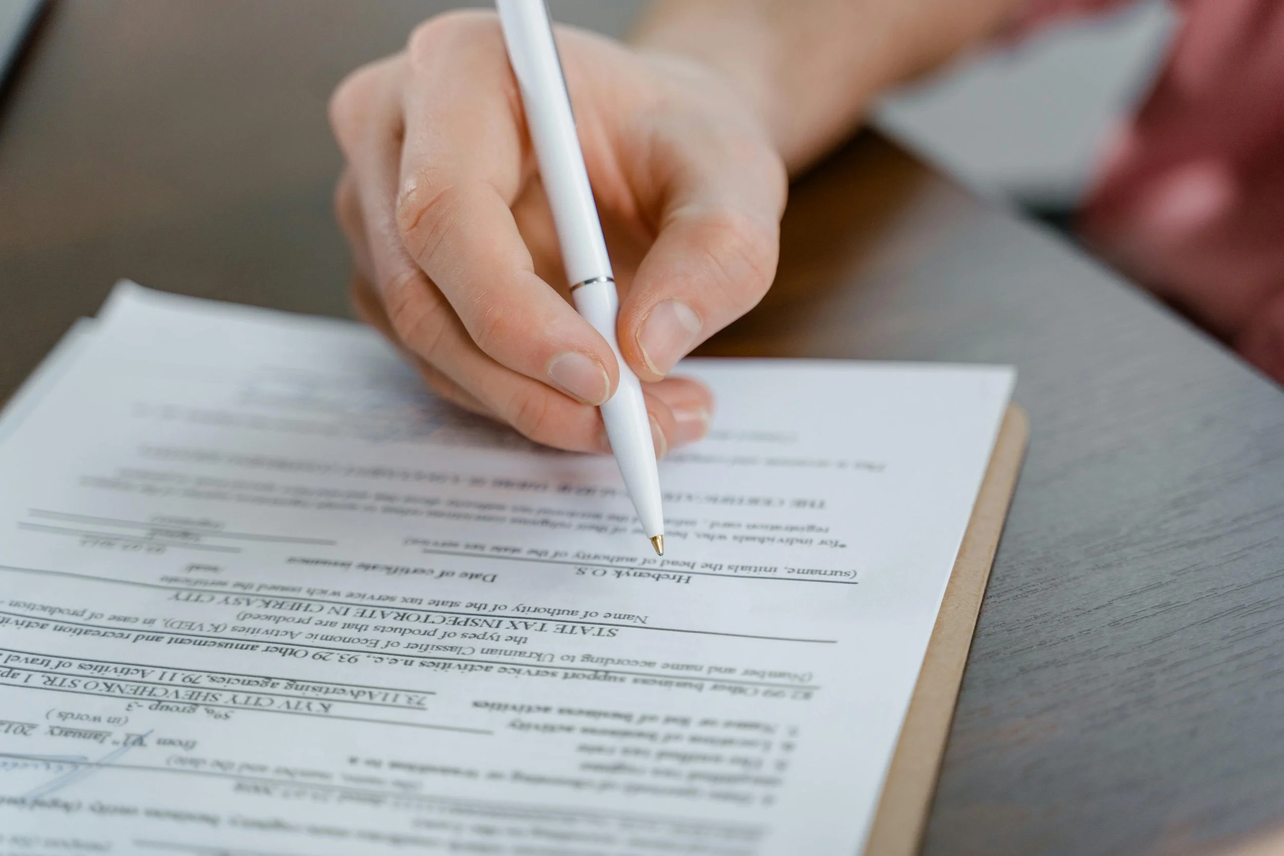 Close-up of a person filling out a form with a white pen on a wooden table.