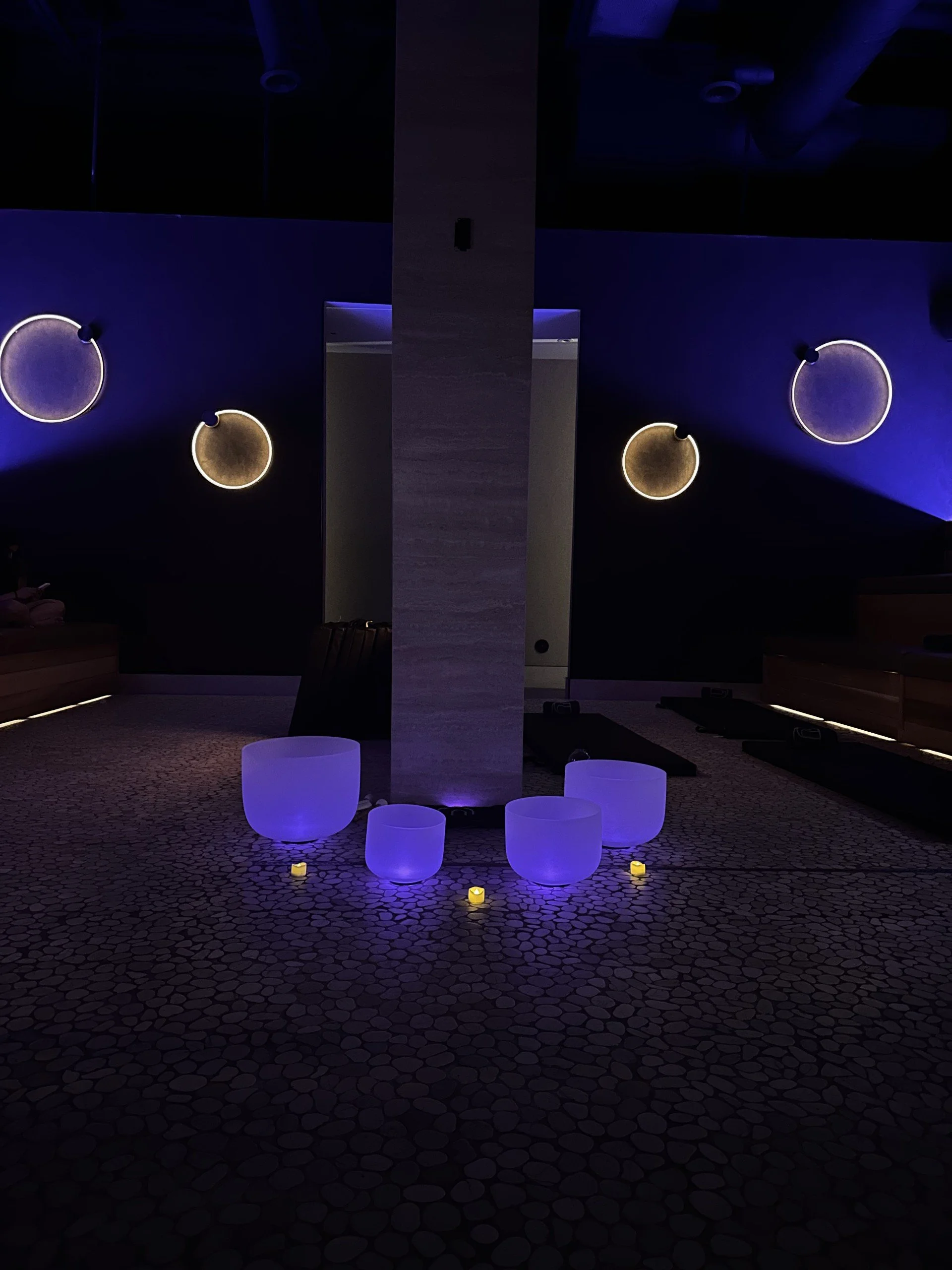 Indoor space with pebble stone floor, decorative white bowls, and yellow glowing candles. Modern circular wall lights on dark walls and a tan central pillar.