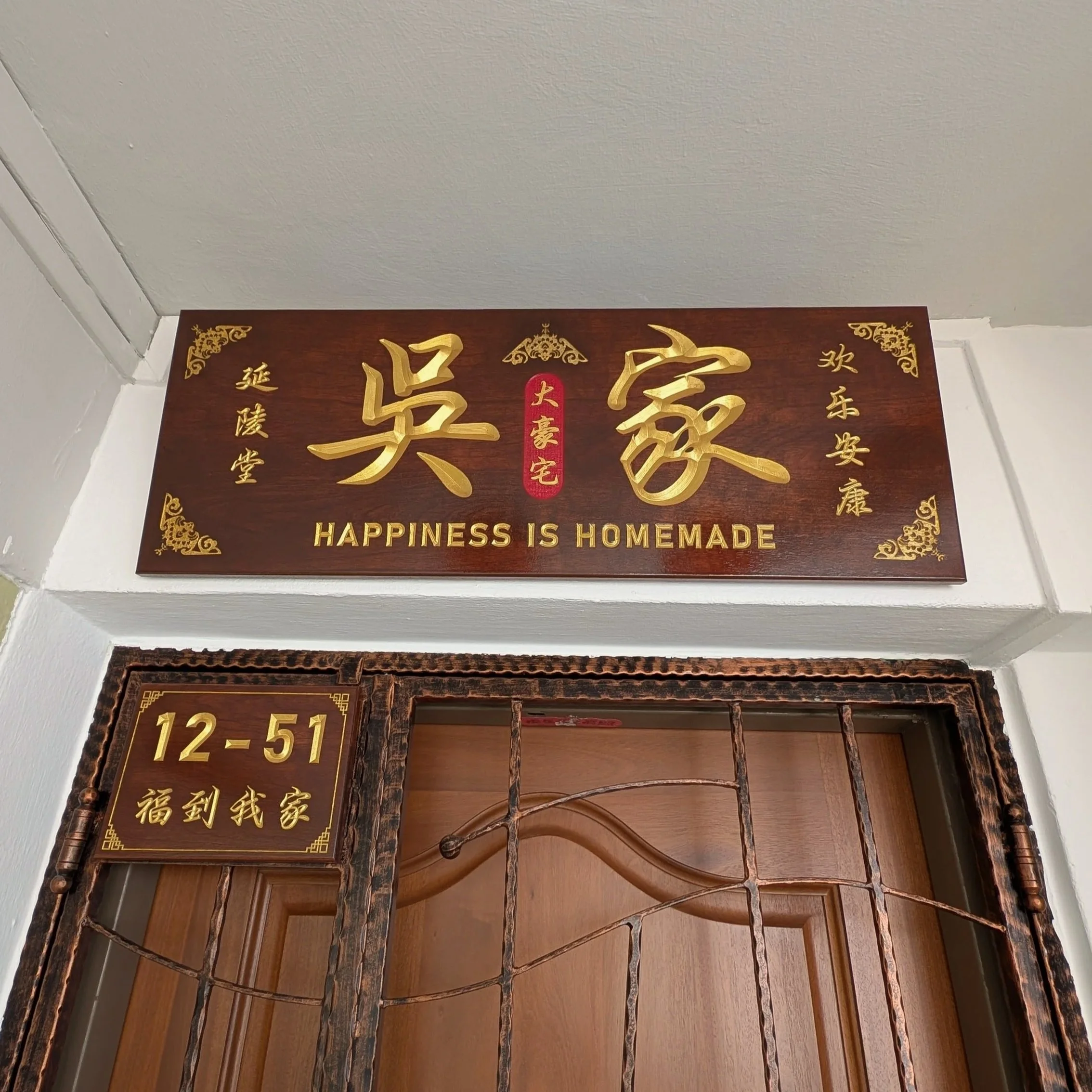Sign above a door with Chinese characters and English text reading 'Happiness is Homemade' on a wooden background, with ornate gold accents and a red stamp in the center.
