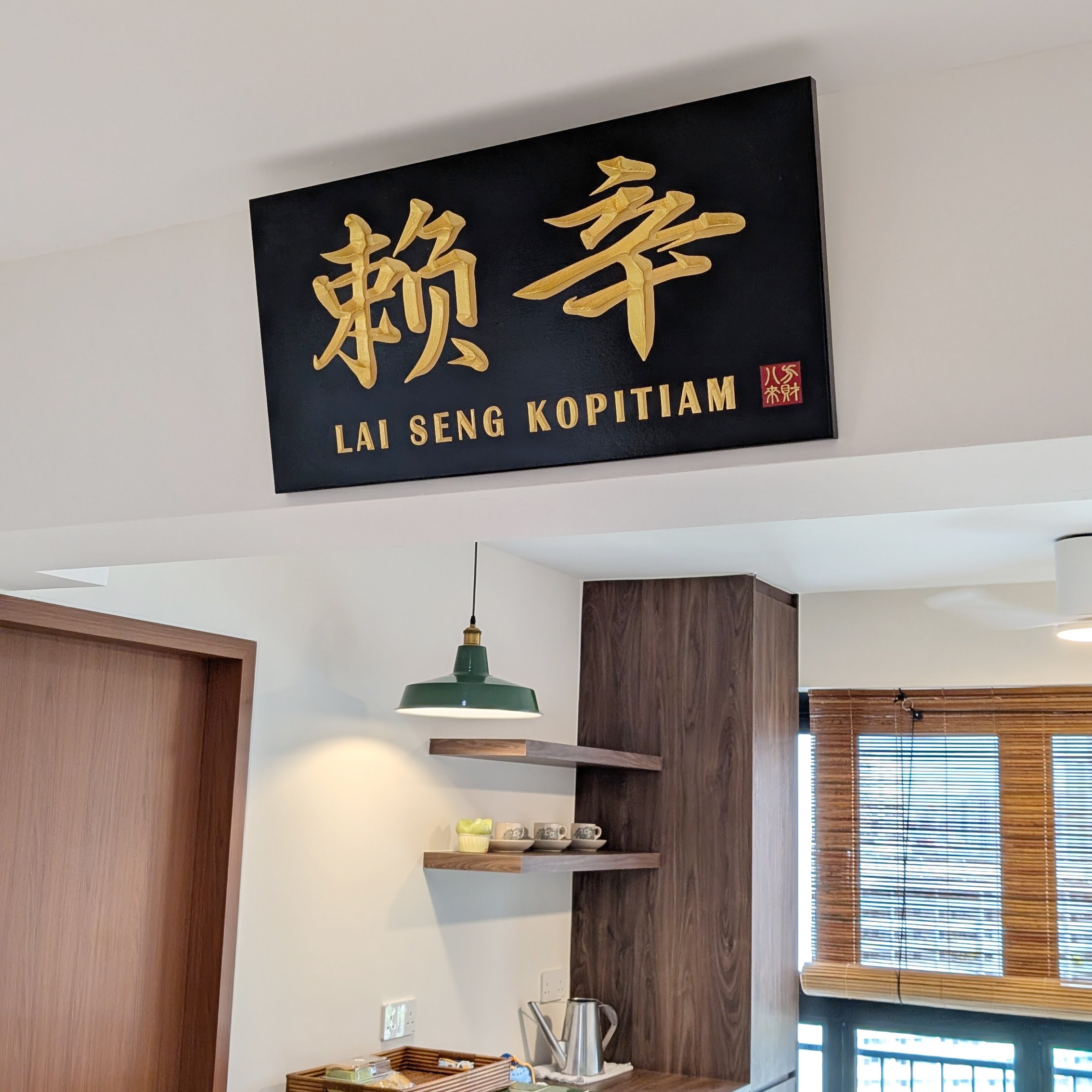 A modern kitchen interior with white walls, wooden cabinets, and black and gold signage reading 'Lai Seng Kopitiam' with Chinese characters. There are hanging green pendant lamps, a shelf with cups and bowls, and a window with blinds.