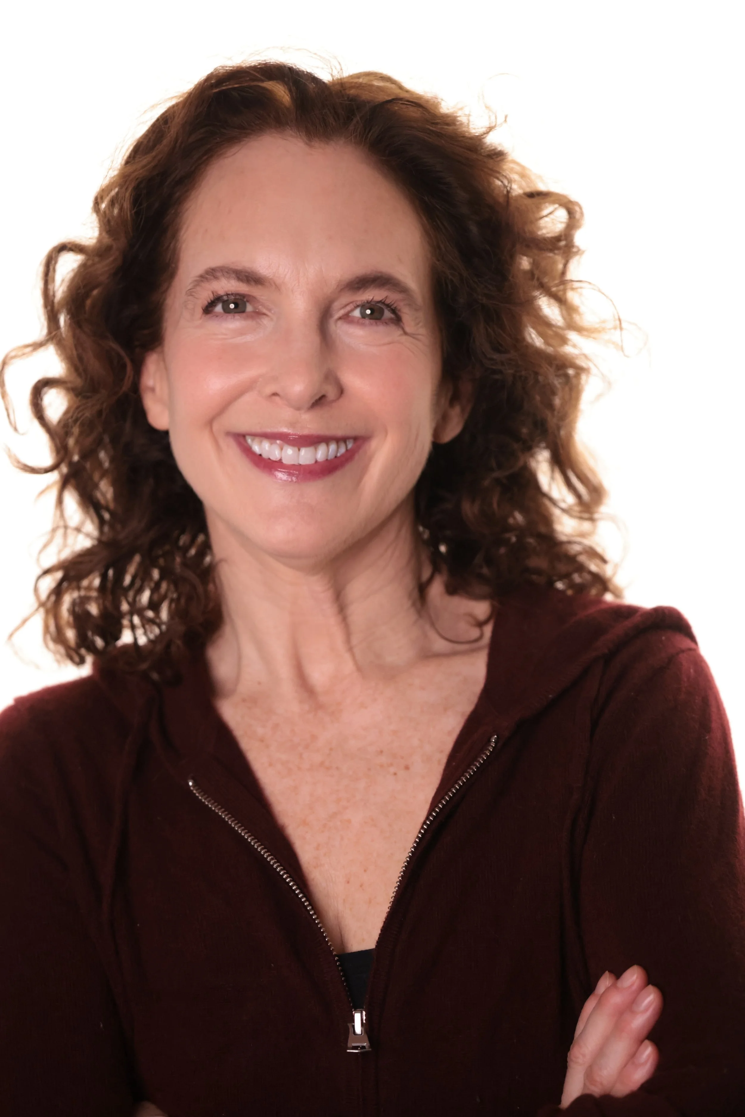 A woman with curly red hair smiling, wearing a dark red zip-up sweater, against a plain white background.