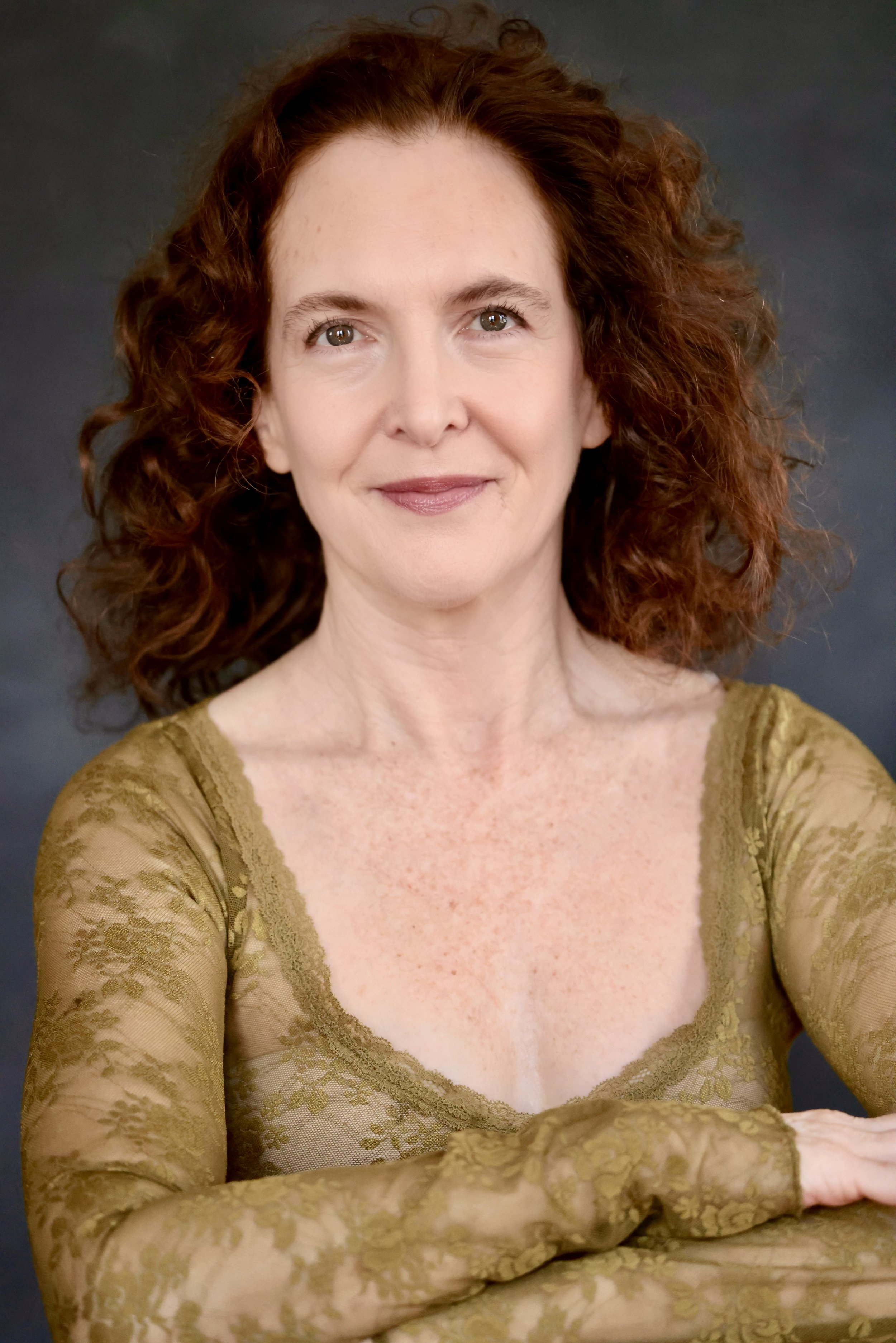A woman with red, curly hair and fair skin, wearing a gold lace long-sleeve top, smiling softly with a dark background.