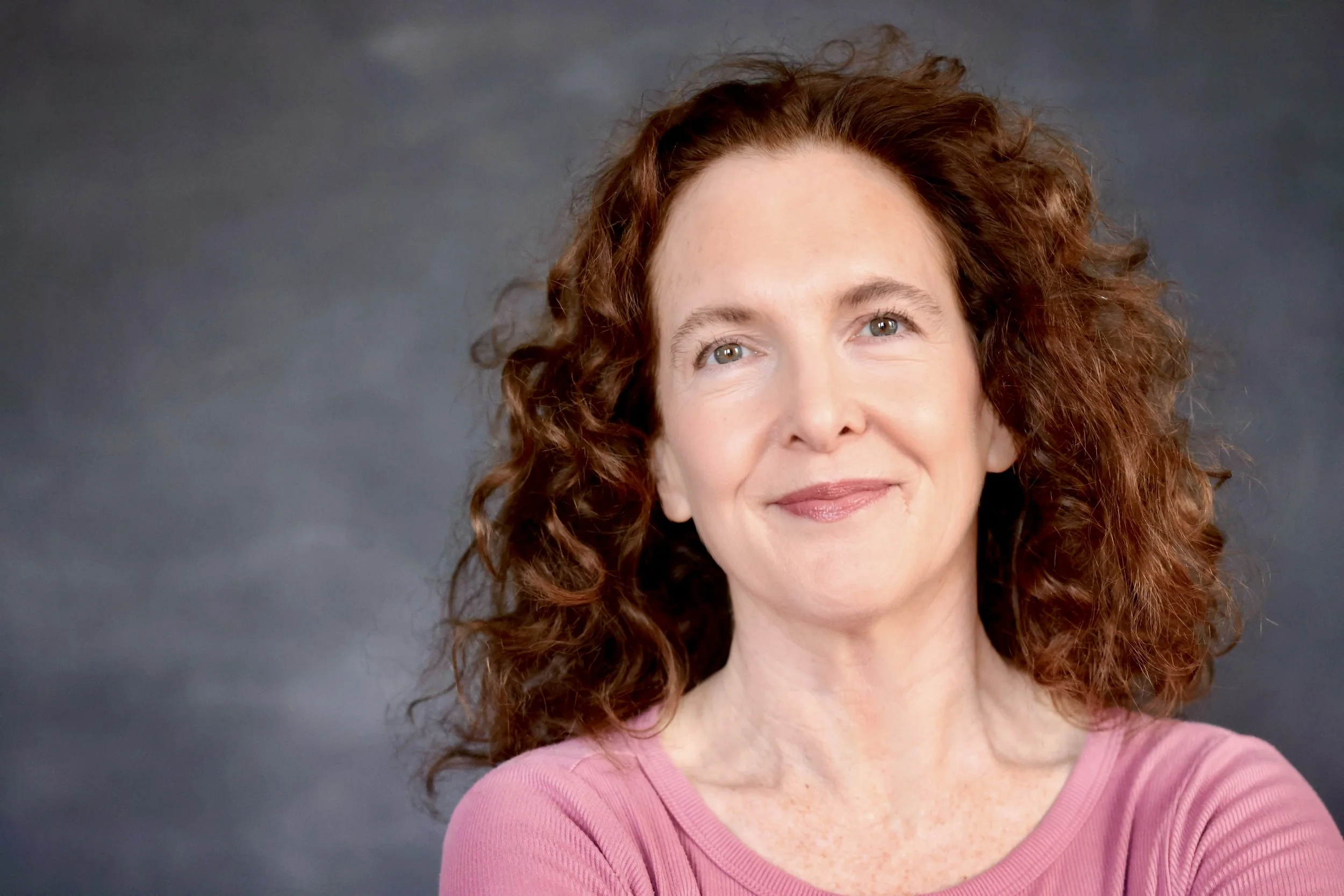 A woman with curly red hair and fair skin smiling, wearing a pink top, against a dark gray background.
