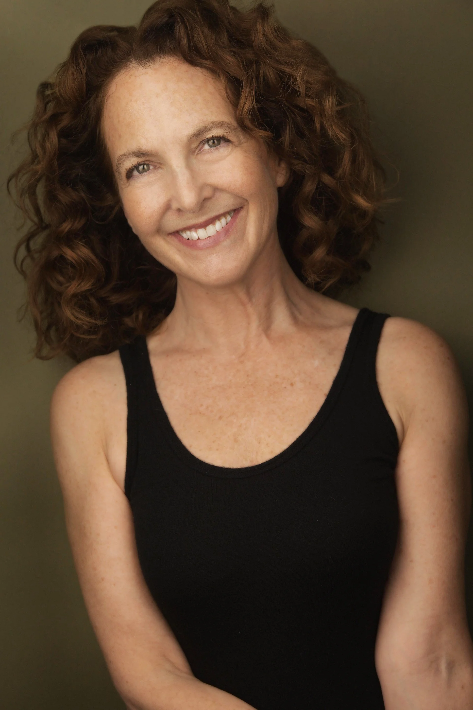 A woman with curly red hair smiling and wearing a black tank top, standing against a plain background.