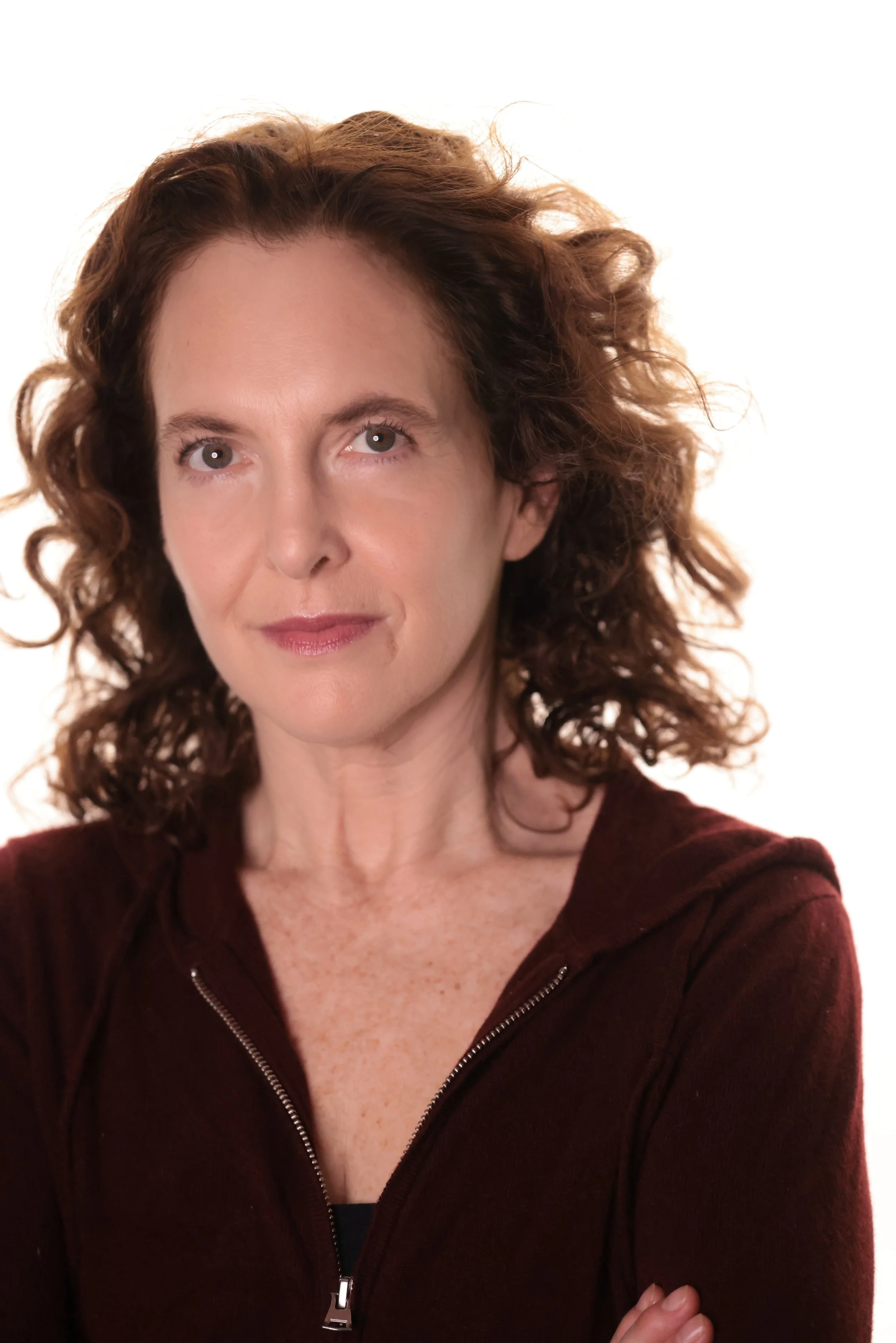 Close-up of a woman with curly brown hair, fair skin, wearing a maroon zip-up jacket, smiling softly, against a white background.