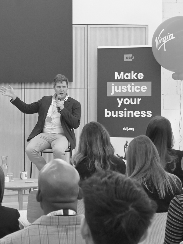 Black and white photo of Virgin Group's Matthias Stausberg presenting on a 2024 W&J Summit panel in London. Behind him is a large balloon with the Virgin logo, and an RBIJ banner that says "make justice your business"