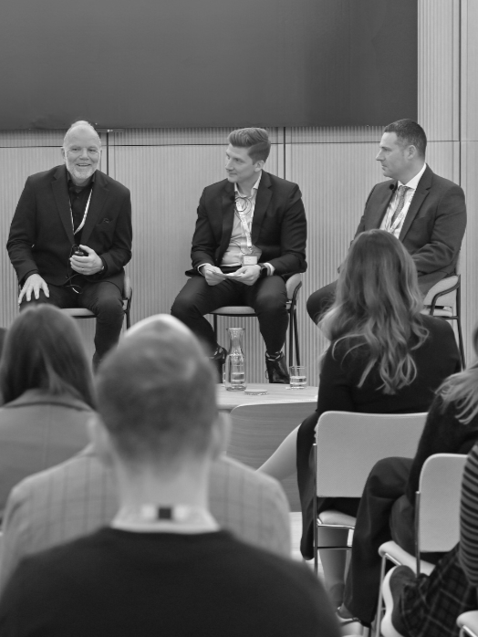 Black and white photo of Iceland Foods' Paul Cowley, SBFM's Colin Shute, and Timpson Group's Darren Burns speaking on a 2024 W&J Summit panel in London.