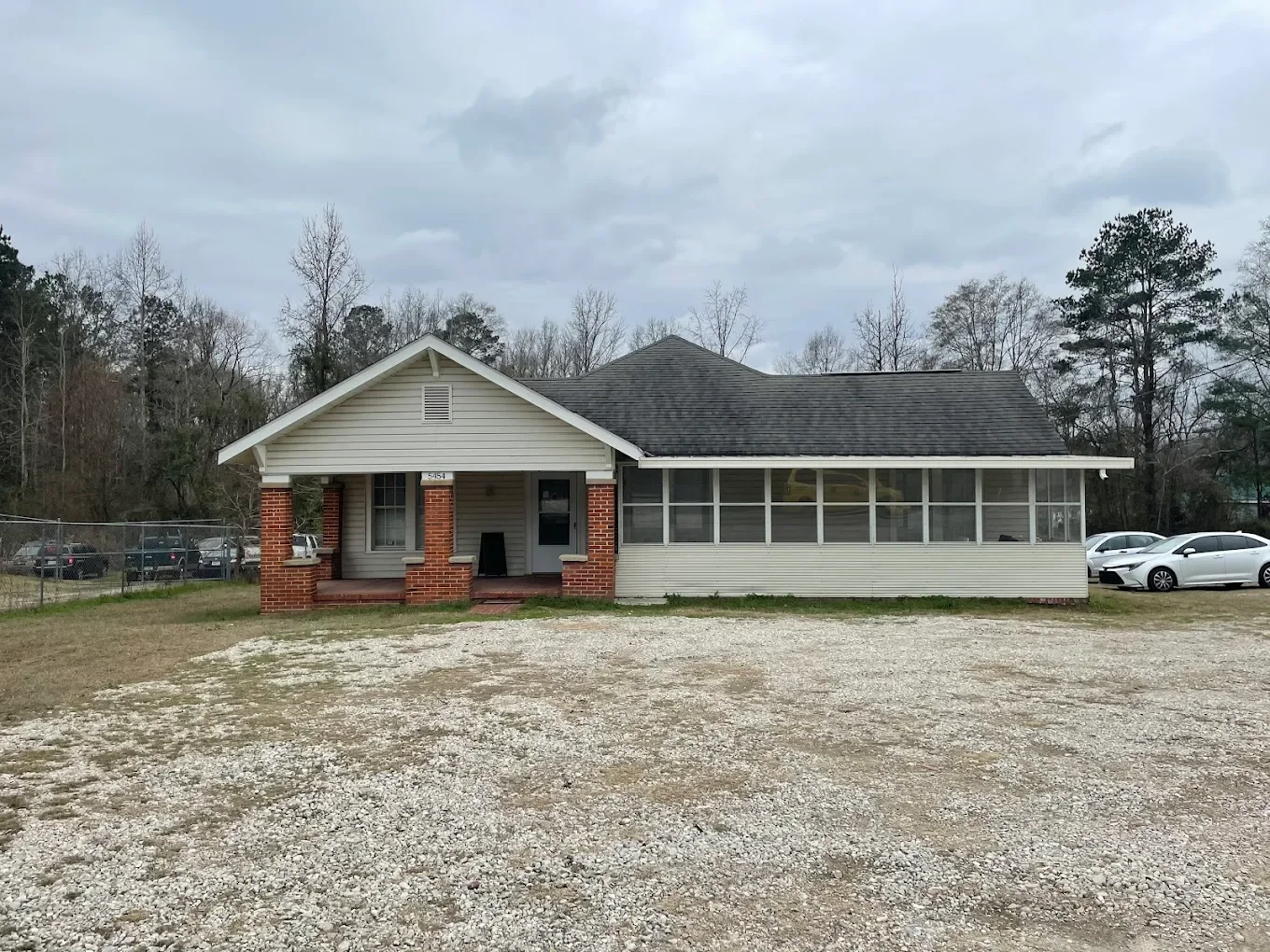 A single-story house with a porch, gray siding, brick columns, and a screened-in patio area, surrounded by a gravel lot and trees in the background.