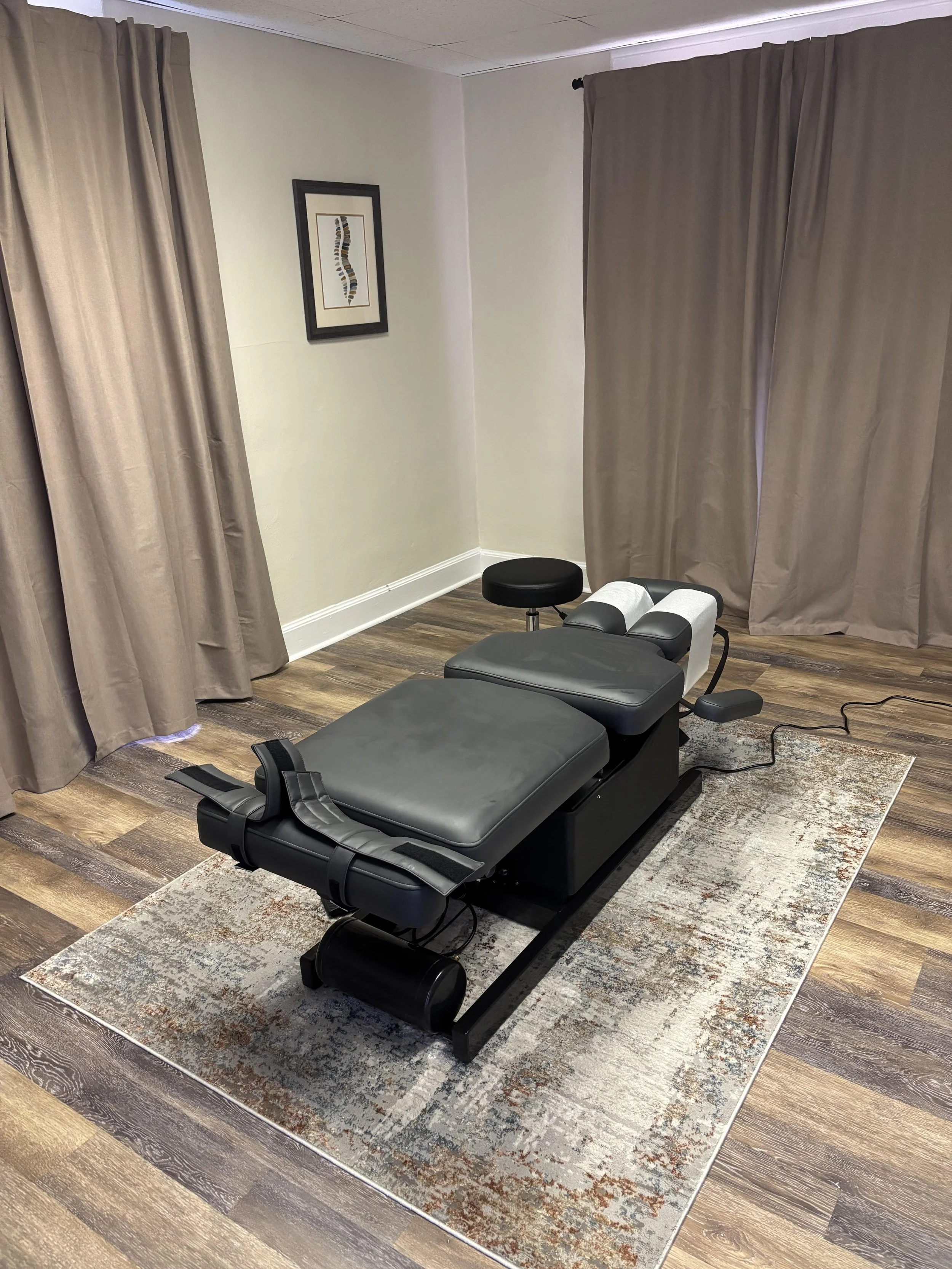 A  chiropractic treatment room with agrey treatment table on a decorative rug, with curtains on the sides and a framed artwork of a colorful spine on the beige wall.