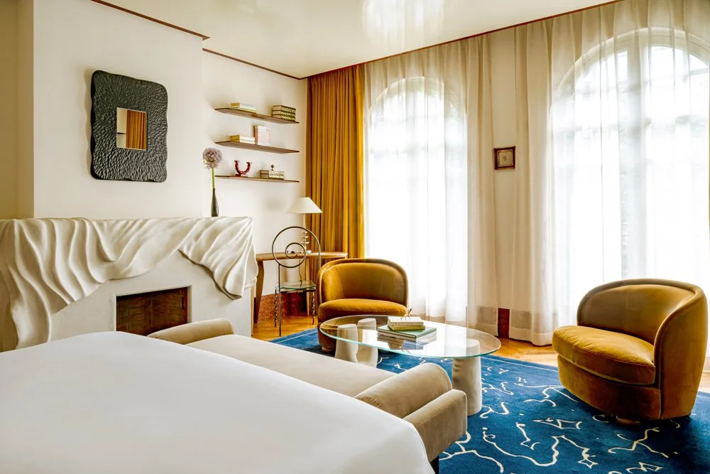 Salon de chambre d'hôtel avec deux fauteuils jaunes, un lit, une table basse en verre avec des livres, une grande fenêtre avec rideaux blancs et dorés, décoration moderne.