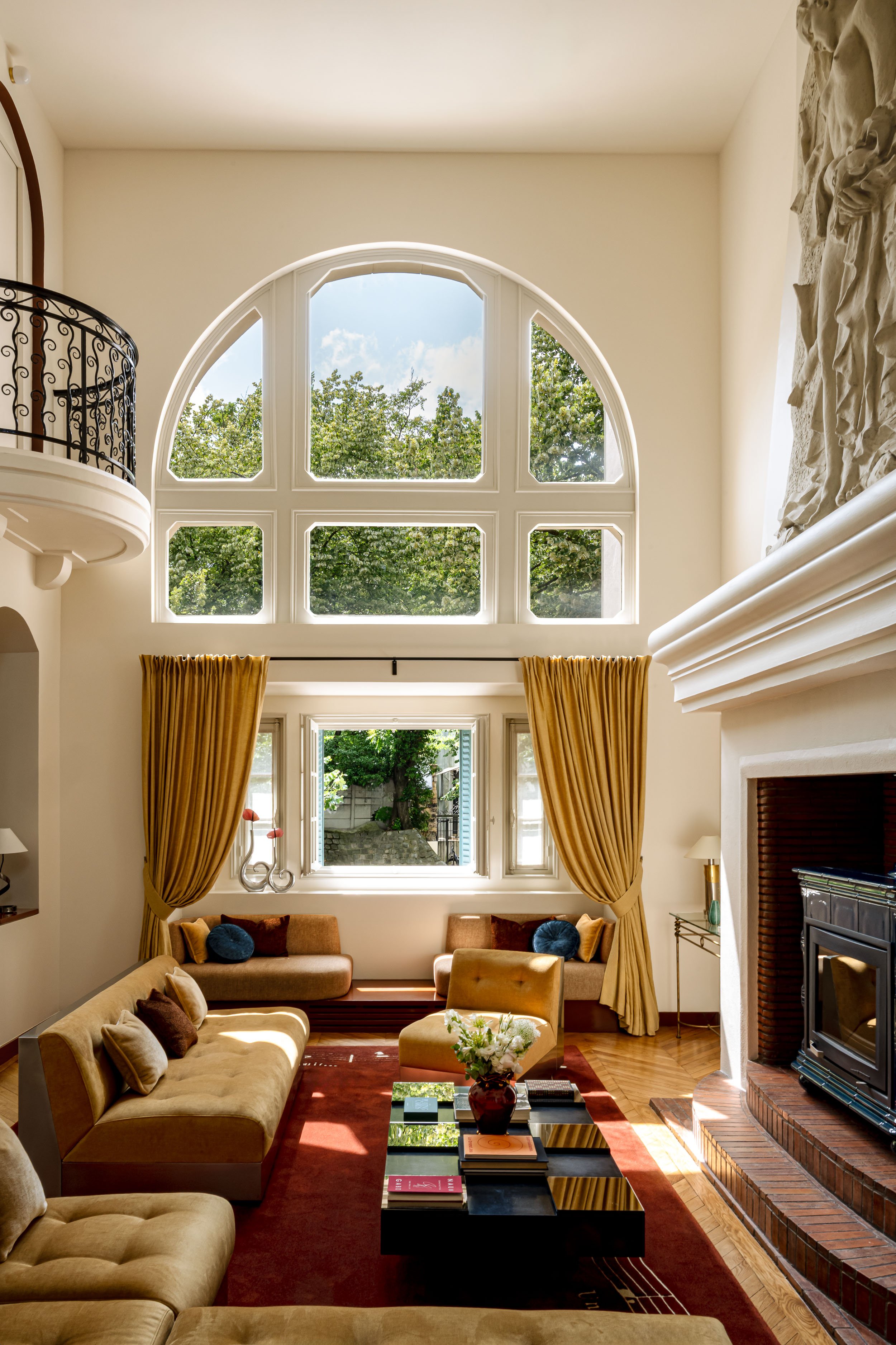 Salon lumineux avec de grandes fenêtres, canapé beige, fauteuils en cuir, cheminée en brique, rideaux dorés, table basse avec livres, décoration florale.