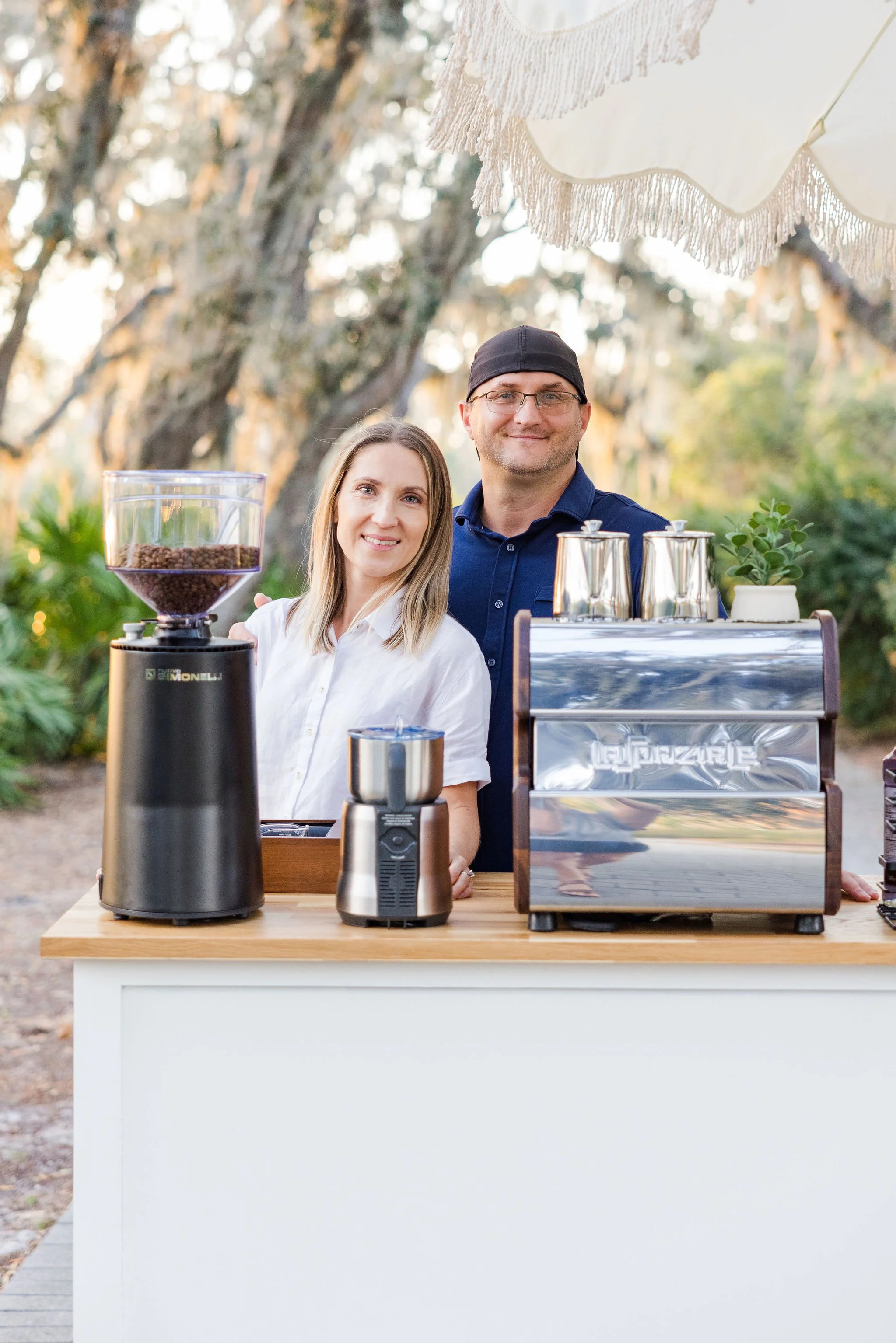 Two people standing behind a coffee bar setup with an espresso machine, coffee grinder, and cups, outdoors.