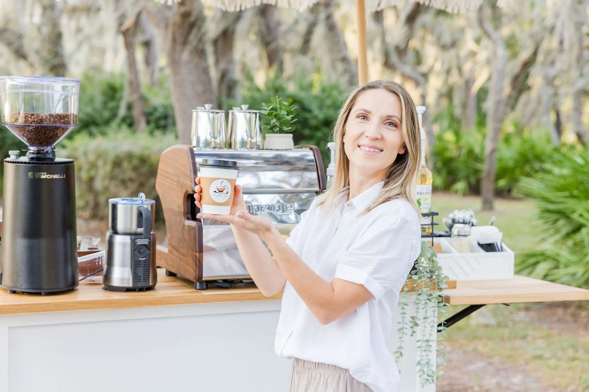 Woman holding coffee cup at outdoor coffee stand, with coffee equipment on a counter, and trees in the background.