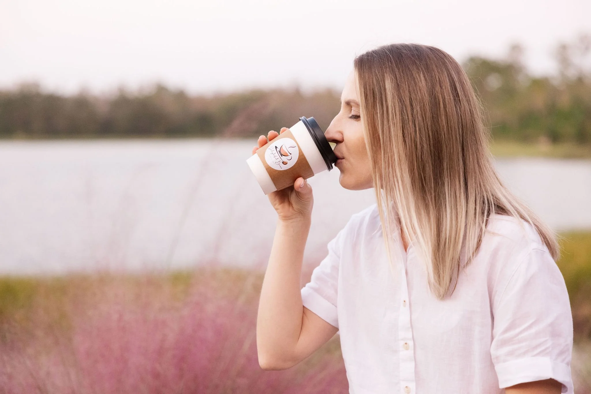 Woman drinking from a to-go coffee cup outdoors, wearing a white shirt, in front of a scenic lake background.