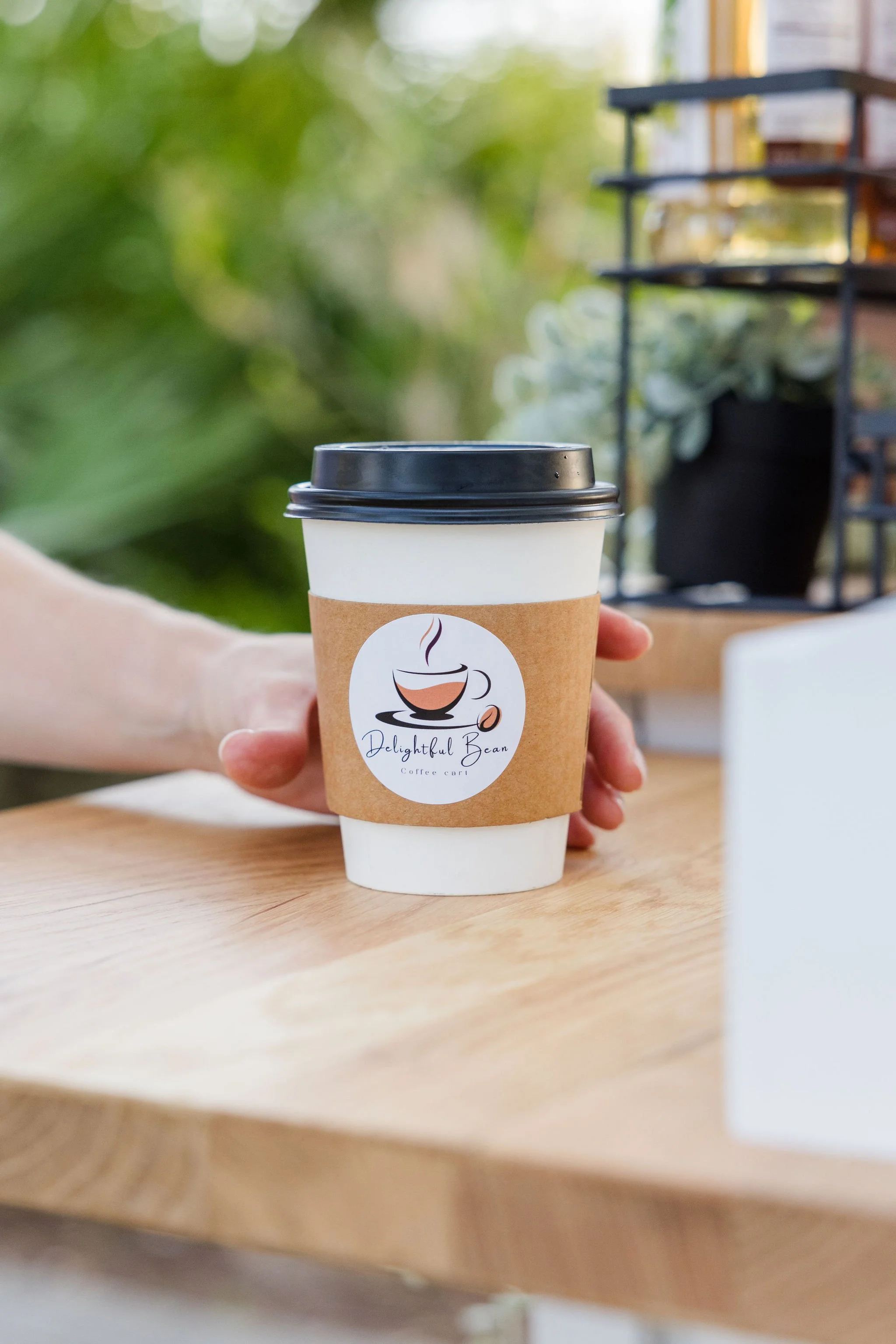 Person holding takeaway coffee cup with logo on wooden table, greenery background.