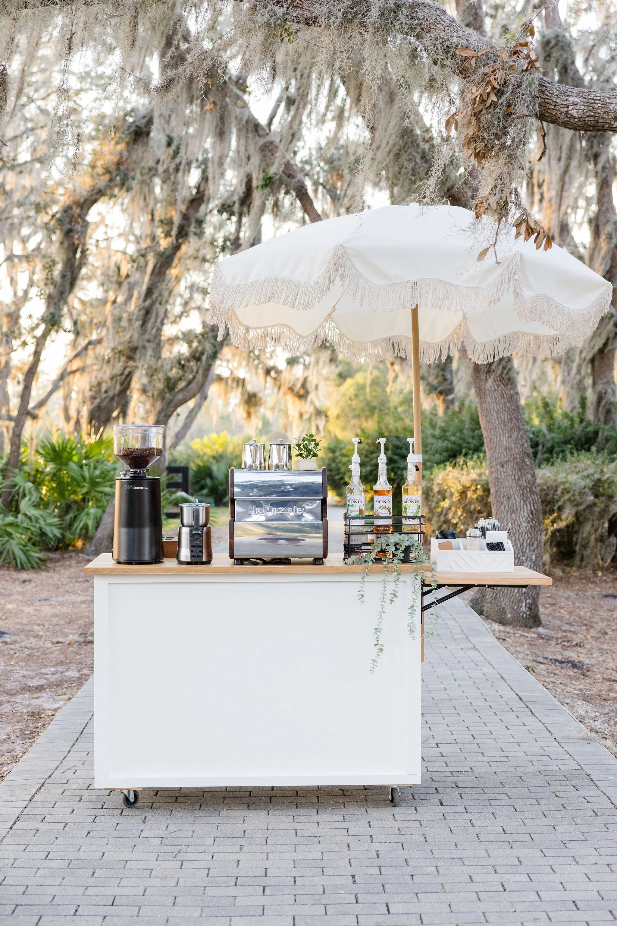 Outdoor coffee cart setup with espresso machine, grinder, coffee syrups, and umbrella.
