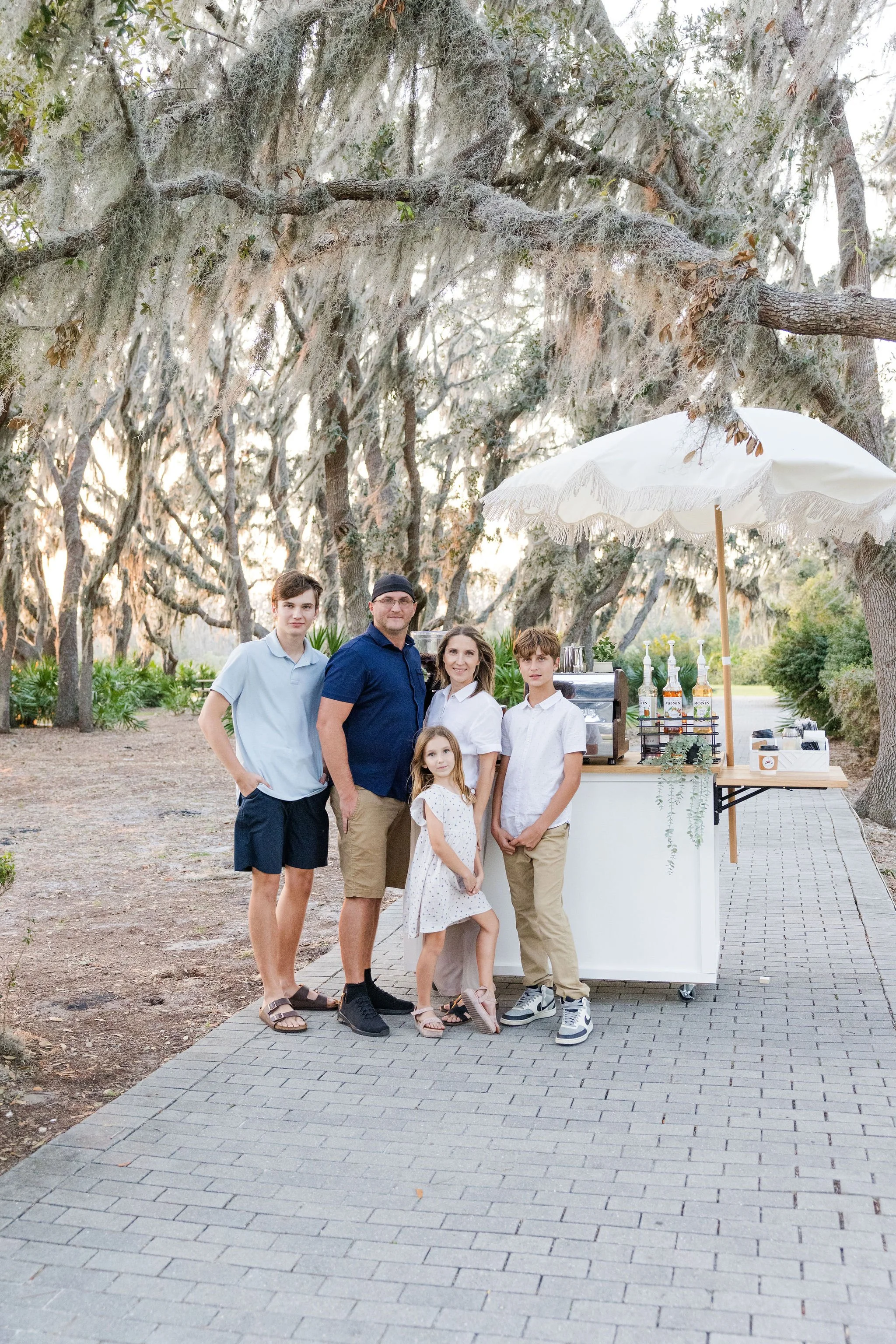 Family posing outdoors in wooded area, near a mobile coffee cart with drinks and umbrella, on a paved path under draped tree branches.