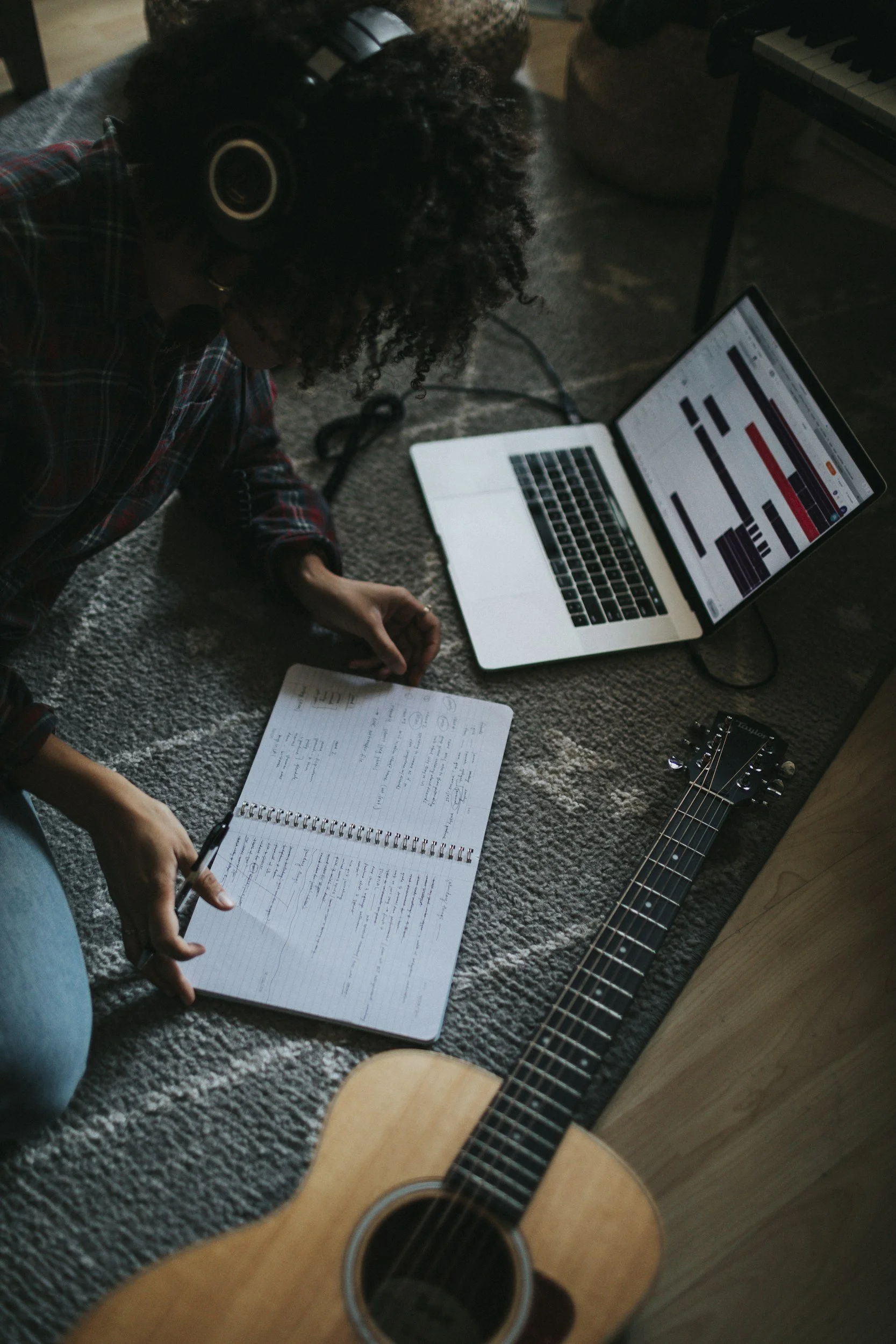 A person lying on a carpeted floor with a notebook, pen, a guitar, and a laptop. The person appears to be writing in the notebook.