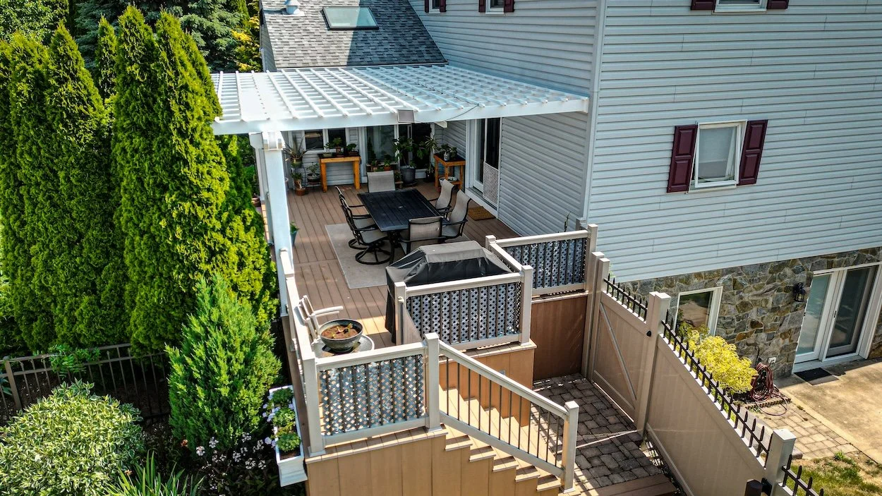Aerial view of a wooden deck with a pergola attached to a house. The deck features outdoor furniture set, several potted plants, and a gas grill. The space is surrounded by tall evergreen trees and a stairway leading to a lower garden area. The house is clad in white siding with a stone accent on the lower level.